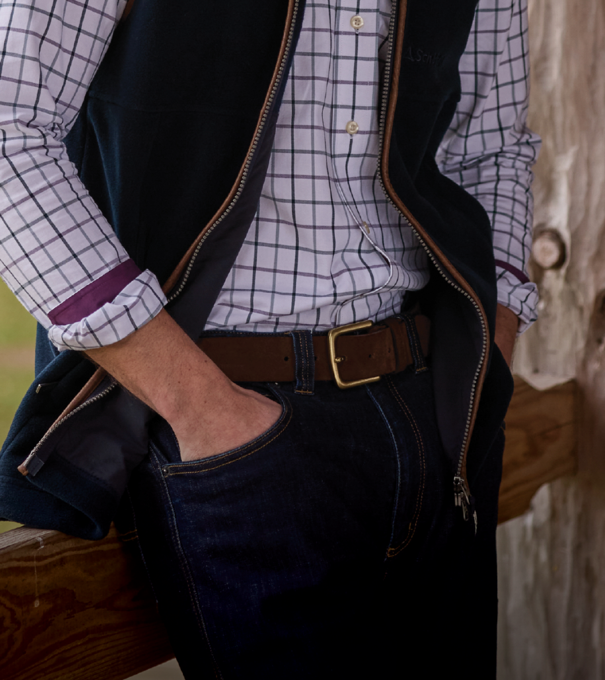 A close-up lifestyle shot of a man wearing a dark navy vest over a white and black checkered shirt, paired with dark blue jeans and a brown leather belt with a gold buckle.