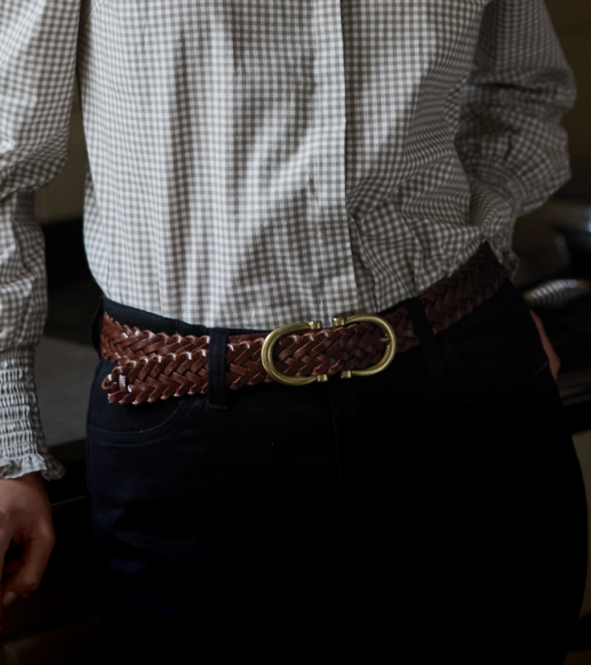 A close-up of a person wearing a brown braided leather belt with a gold-tone oval buckle, paired with a black-and-white gingham shirt and dark trousers.