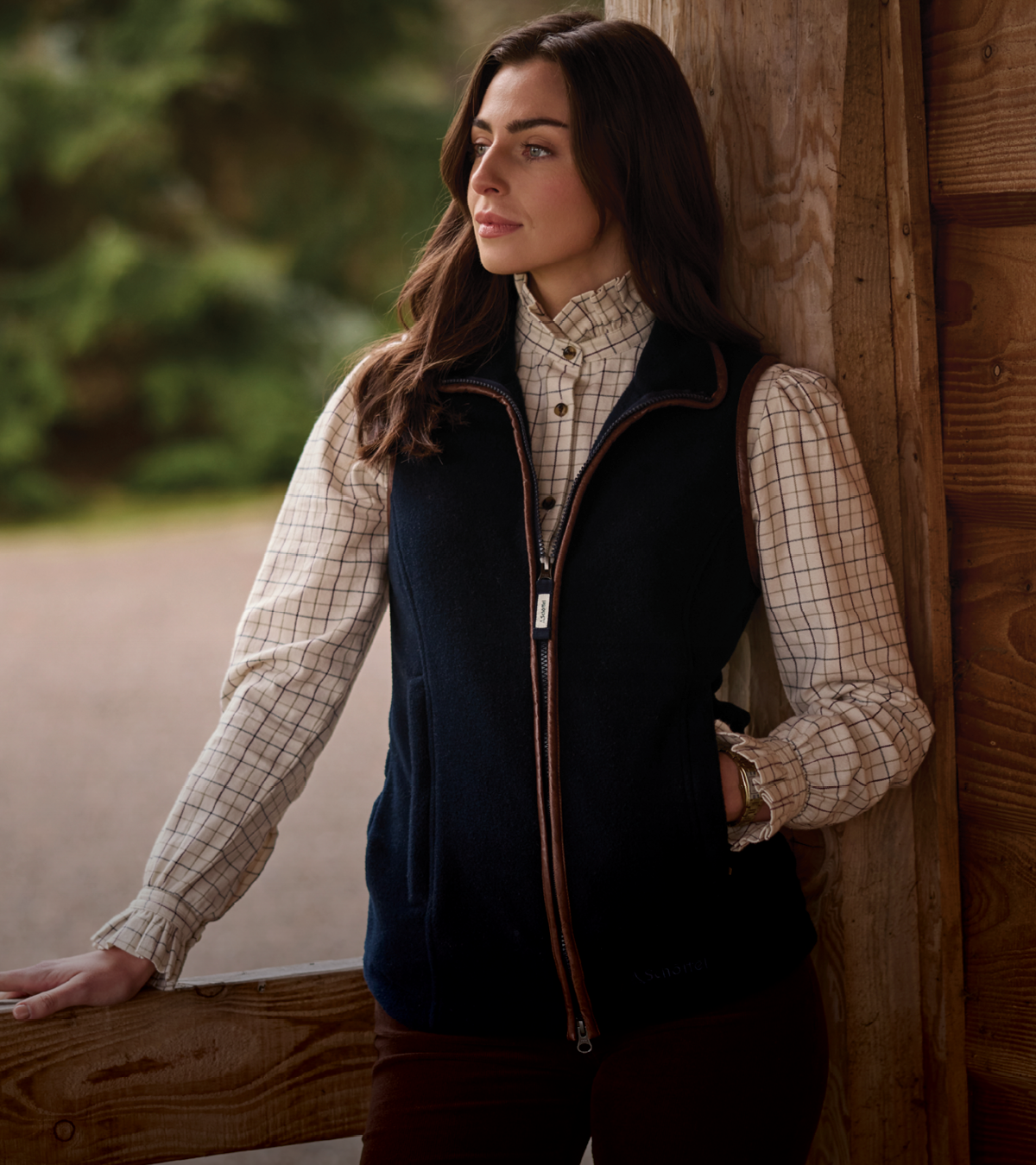 A woman leans against a rustic wooden post wearing a navy blue zip-up fleece gilet over a beige plaid button-down shirt, with a blurred outdoor background.