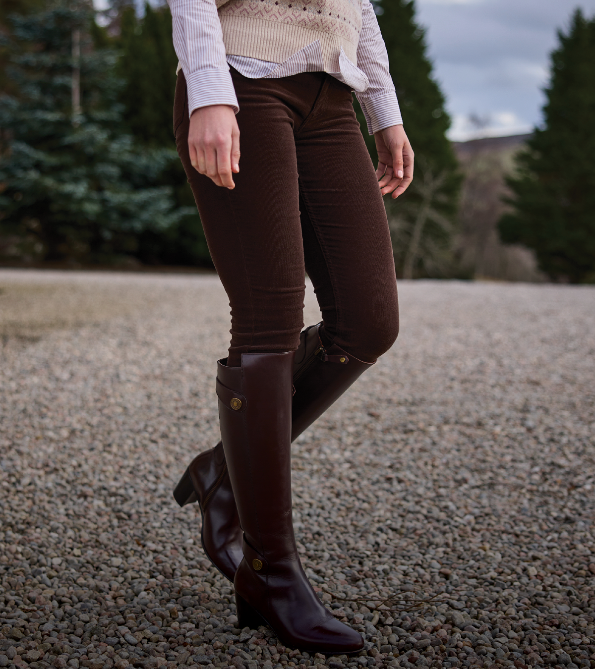 A person outdoors wearing dark brown skinny trousers and matching dark brown knee-high riding boots, standing on a gravel surface with trees in the background.
