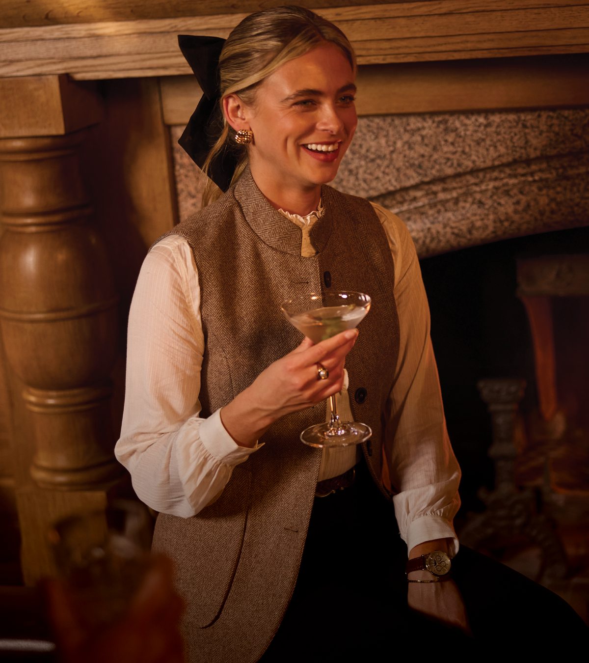 A woman in a warm, dimly lit indoor setting holds a martini glass. She is wearing a brown tweed waistcoat over a white blouse with a high neck and a black hair bow.