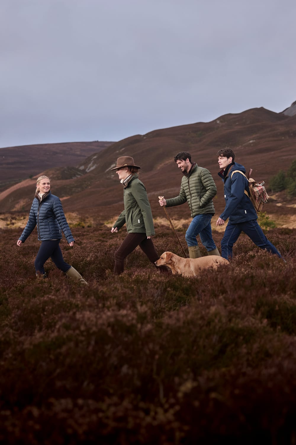 Four people walking outdoors across a moor with hills in the background under an overcast sky. Two of the walkers are wearing puffer jackets, and a golden dog is on a leash in the foreground.