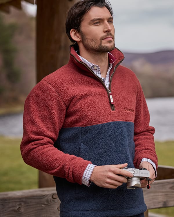A man wearing a two-tone fleece pullover fleece with a red upper section and a navy blue lower section, standing outdoors holding a camera.
