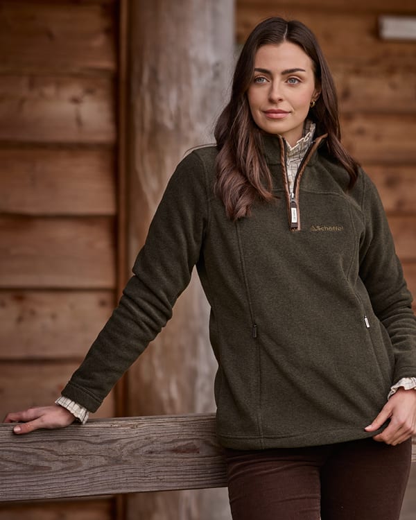 A woman wearing an olive green quarter-zip fleece, leaning against a rustic wooden fence in front of a wooden cabin wall.
