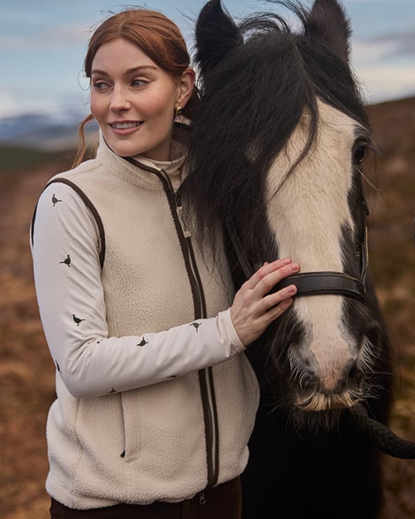 A woman in a cream fleece gilet with brown trim stands outdoors next to a black and white horse, touching its face.