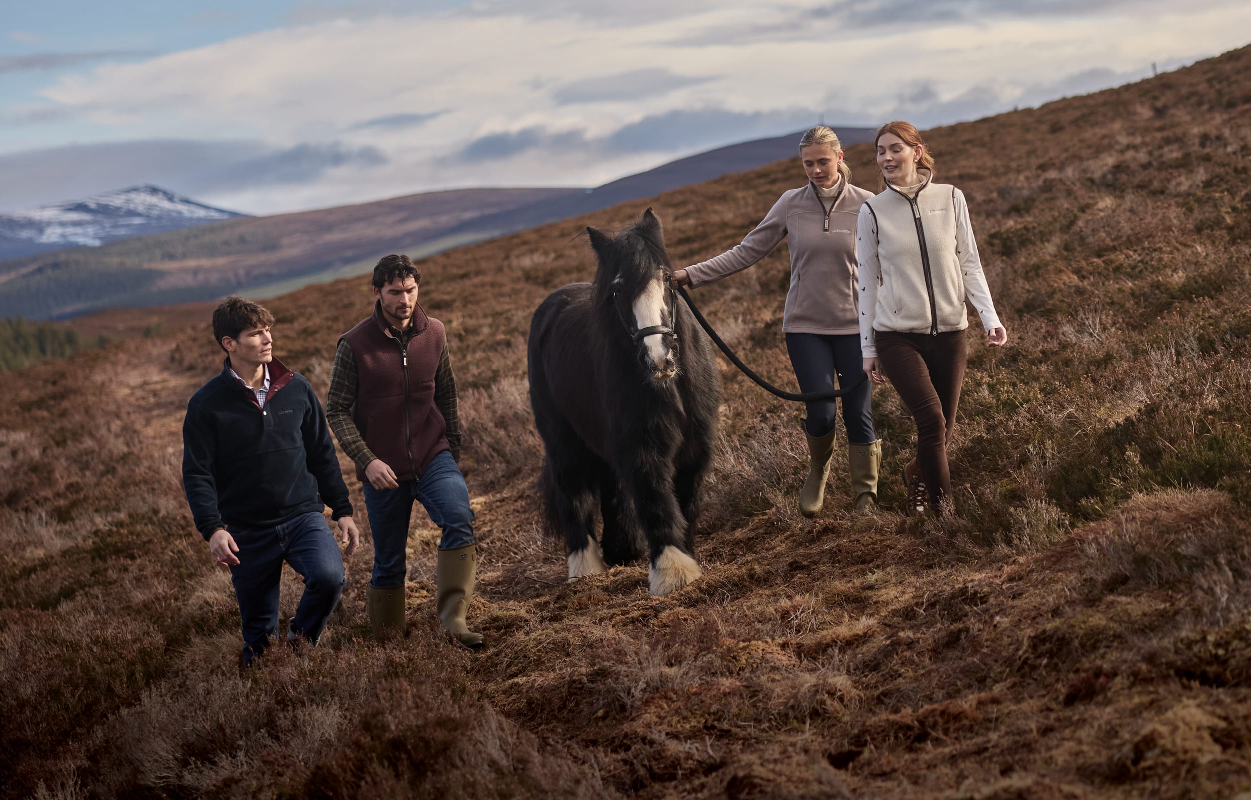 Four people walking with a dark horse on a grassy hillside, wearing outdoor casual jackets and boots in a natural mountain setting.