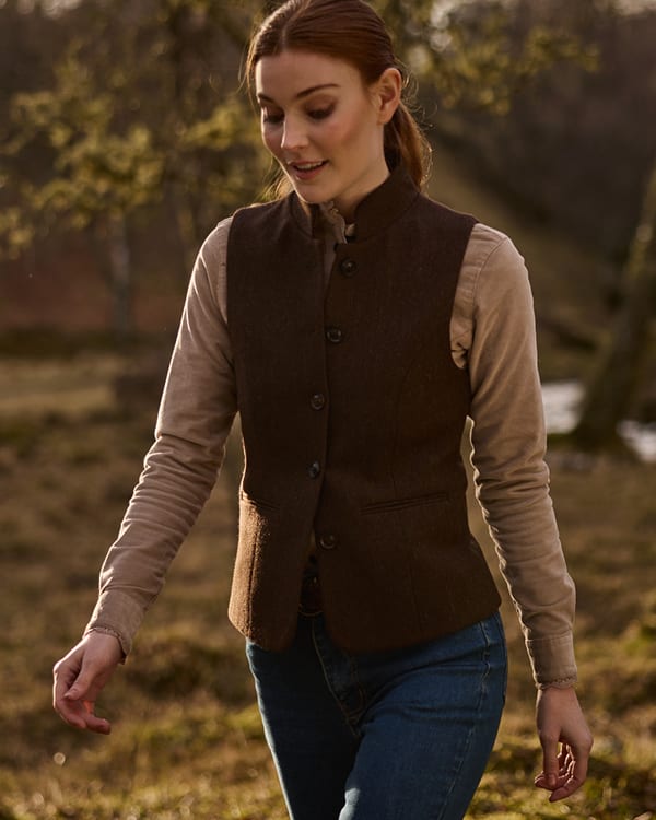 A woman wearing a dark brown wool waistcoat over a beige long-sleeve shirt and blue jeans, walking outdoors in a sunlit field with trees in the background.