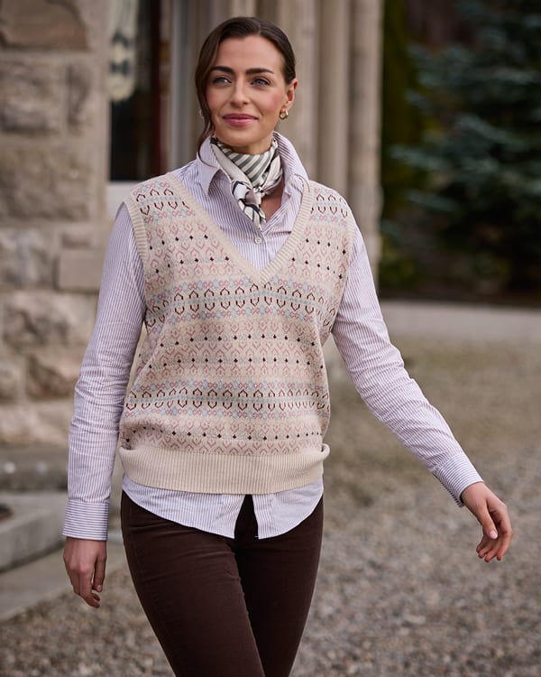 A woman wearing a cream fairisle knit sweater vest over a light pink striped button-down shirt, accessorized with a striped neck scarf and dark brown pants, standing outdoors on a gravel path.