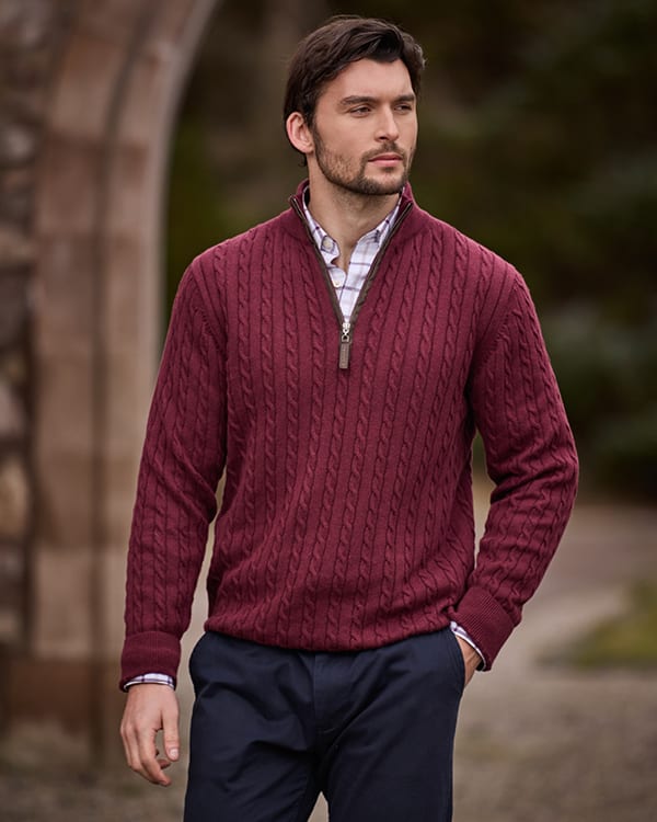 A man wearing a burgundy cable-knit quarter-zip jumper over a white checked shirt and navy trousers, standing outdoors in front of a blurred stone archway.