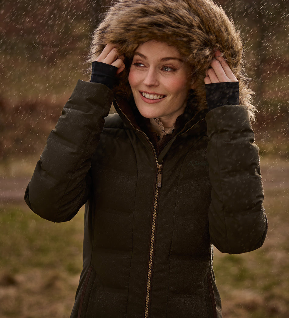 A woman wearing a dark green puffer jacket with a large brown faux-fur hood, standing outdoors in falling rain.