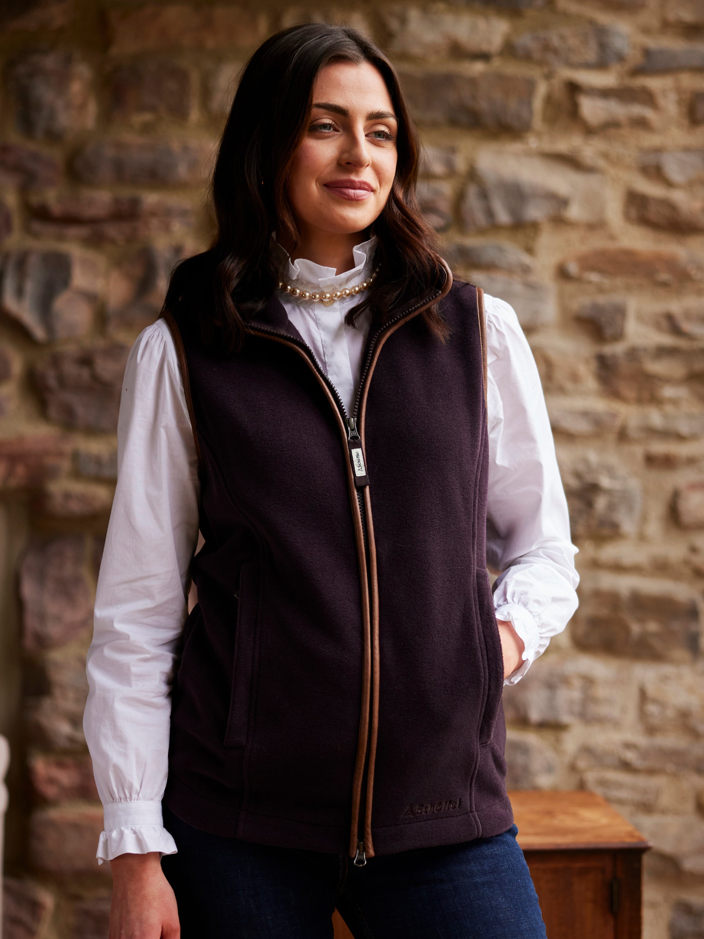 A woman standing in a cottage style house, she is wearing a white shirt, blue trousers and a dark purple gilet.