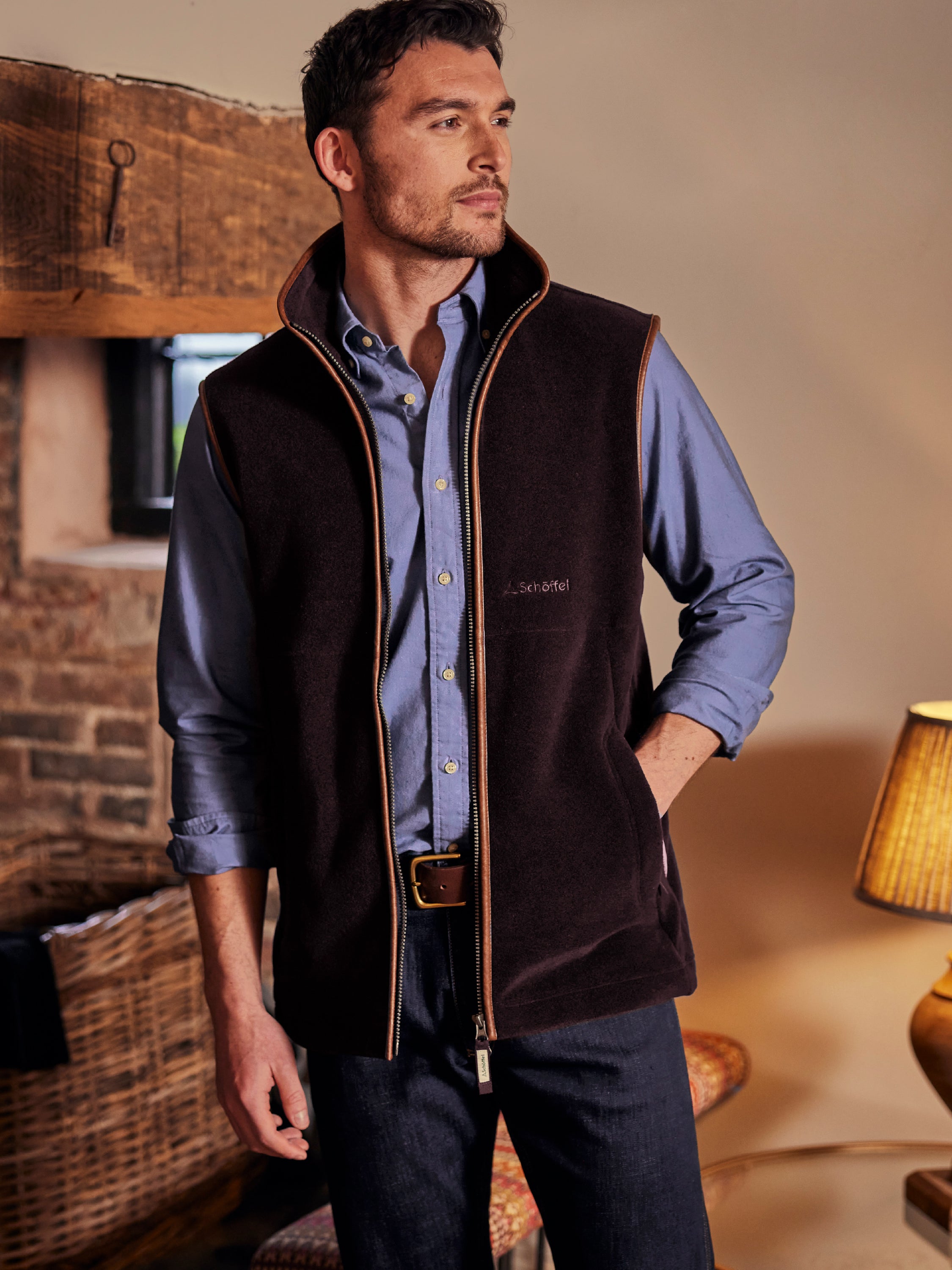 A man standing in a cottage style house. He is wearing dark navy jeans, a light blue shirt and a dark purple gilet.