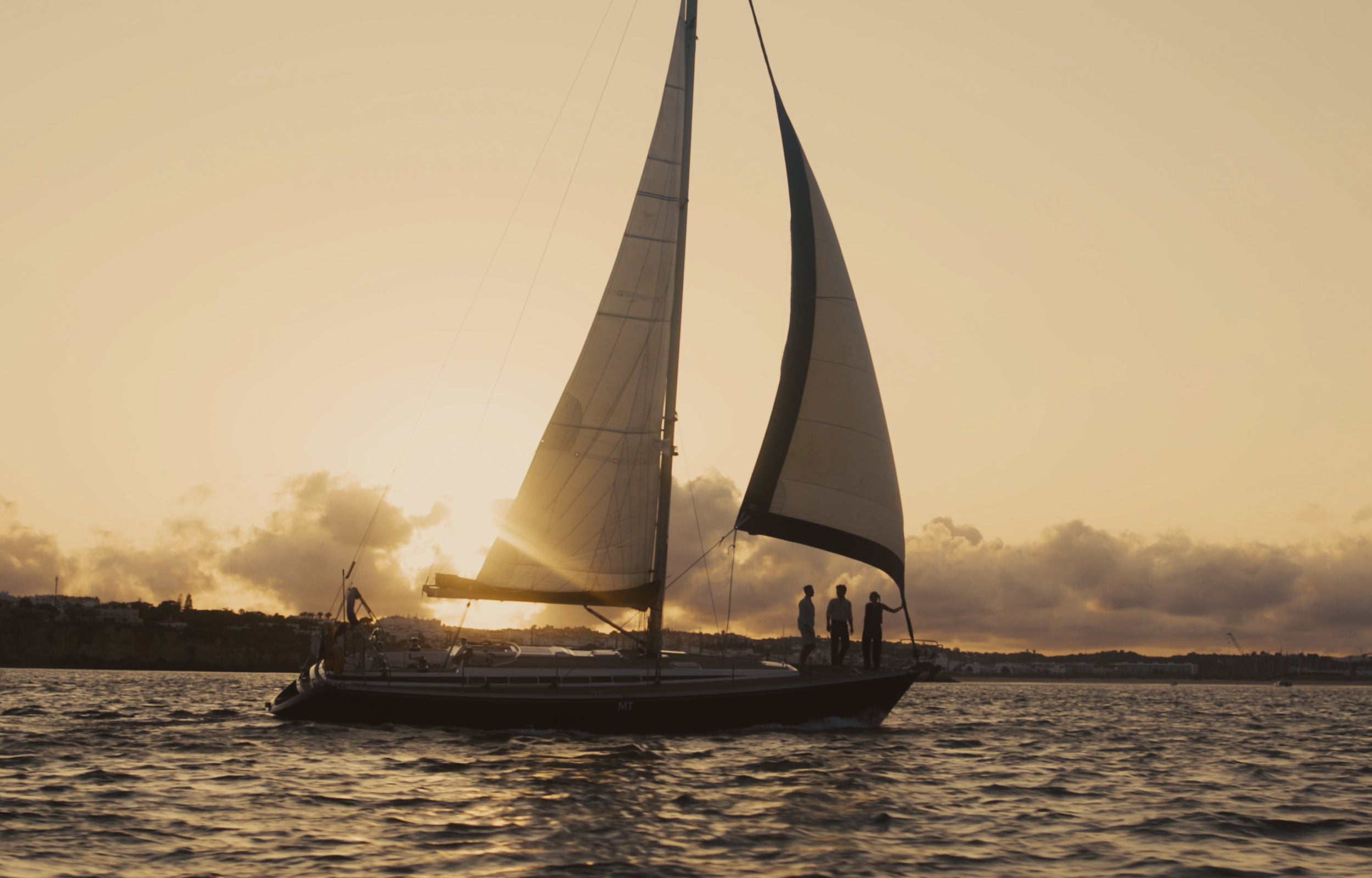 A sailboat on open water at sunset, with the warm golden sun low over the horizon and silhouetted figures standing on the deck.