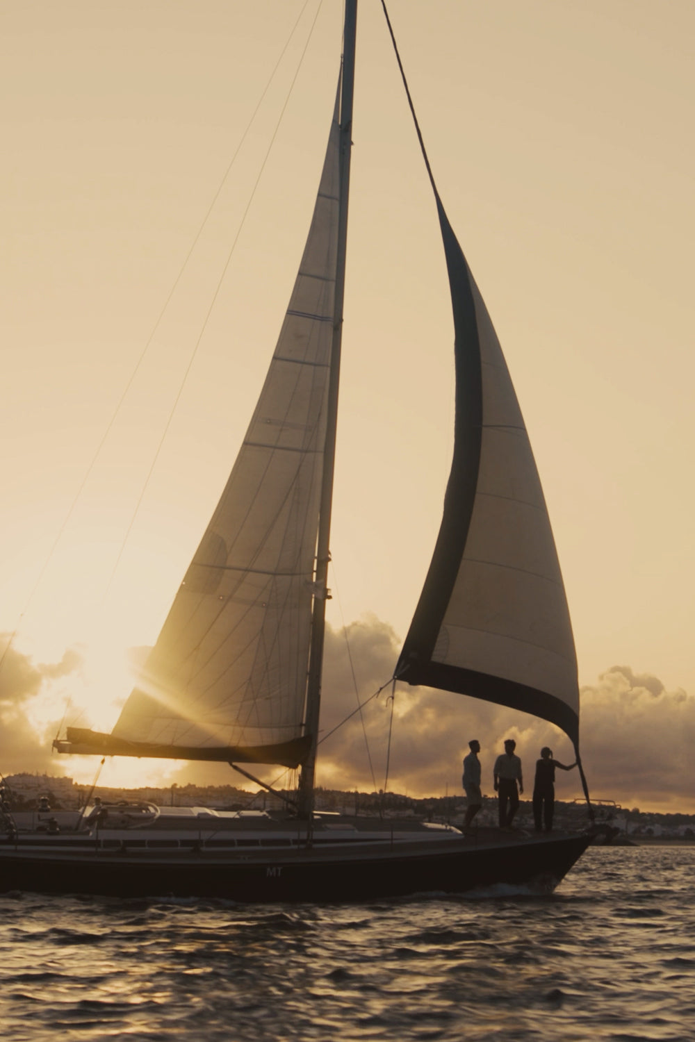 A sailboat on the water at sunset with three people standing on deck, silhouetted against a warm golden sky.