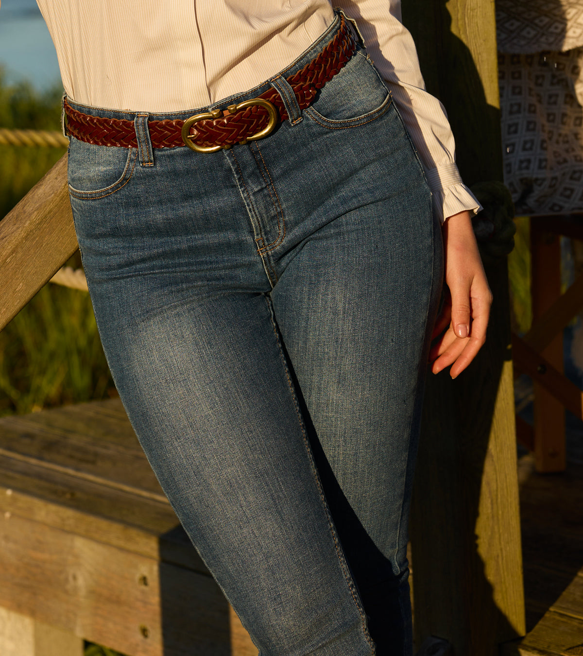 Person wearing blue jeans with a braided belt on a wooden deck.
