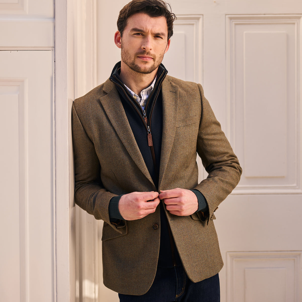 Man wearing a brown blazer over a dark shirt and jeans, standing against a light-colored wall.