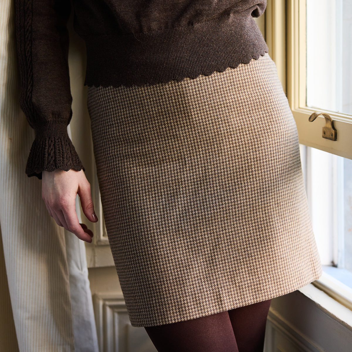 Person wearing a brown sweater and tweed pattern skirt standing by a window.