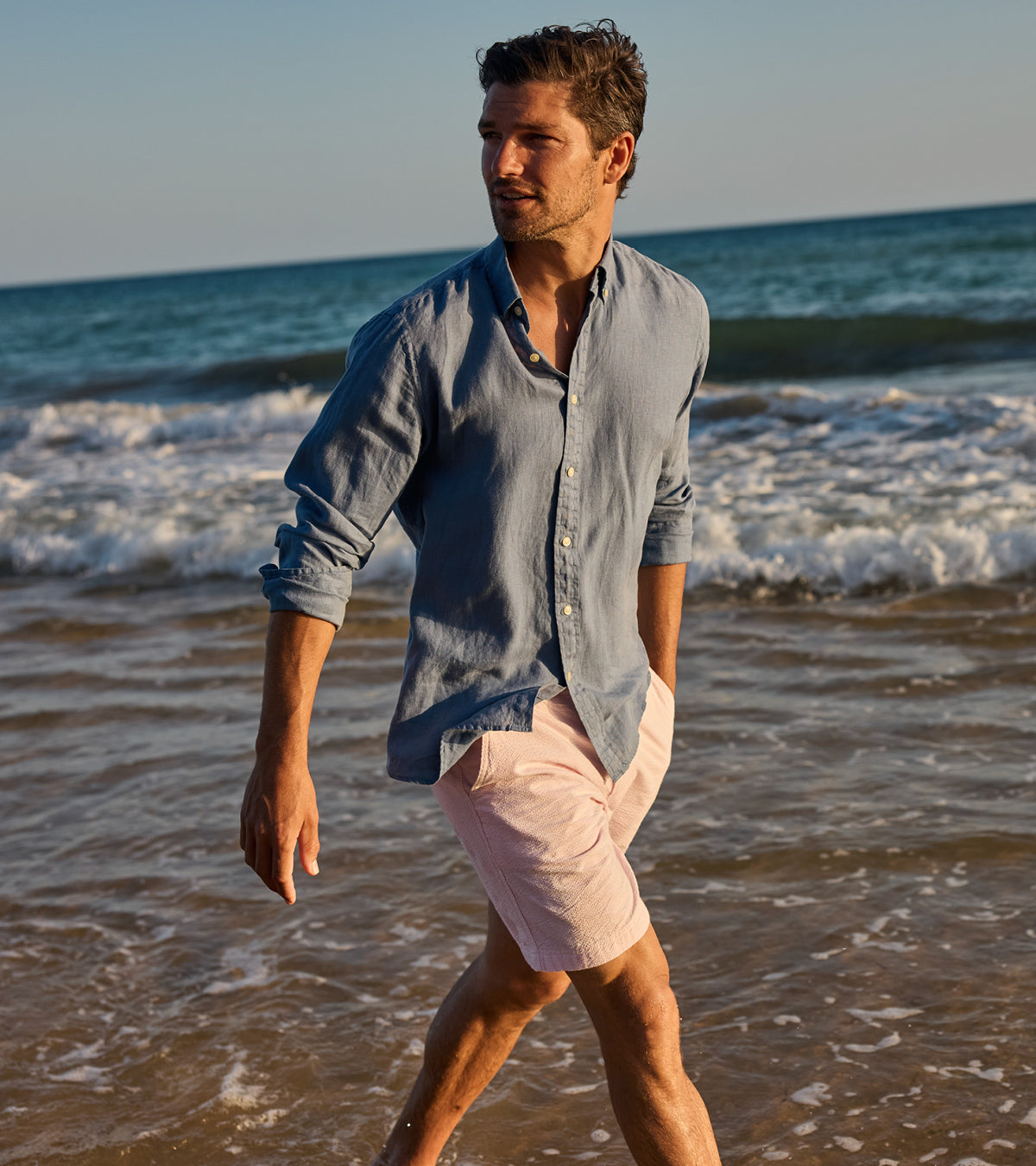 Man walking on a beach wearing a light blue shirt and pink shorts.
