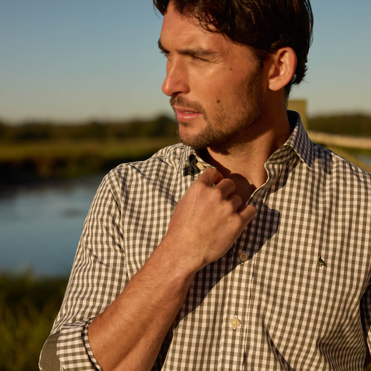 Man adjusting his checkered shirt outdoors with a blurred natural background