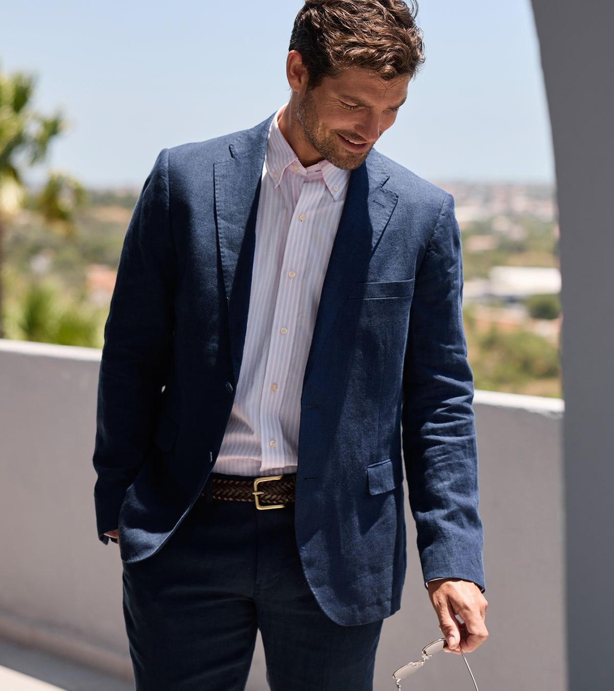 A man wearing a navy blue linen blazer over a light pink and white striped dress shirt, paired with navy trousers and a brown belt, standing outdoors on a sunny day.