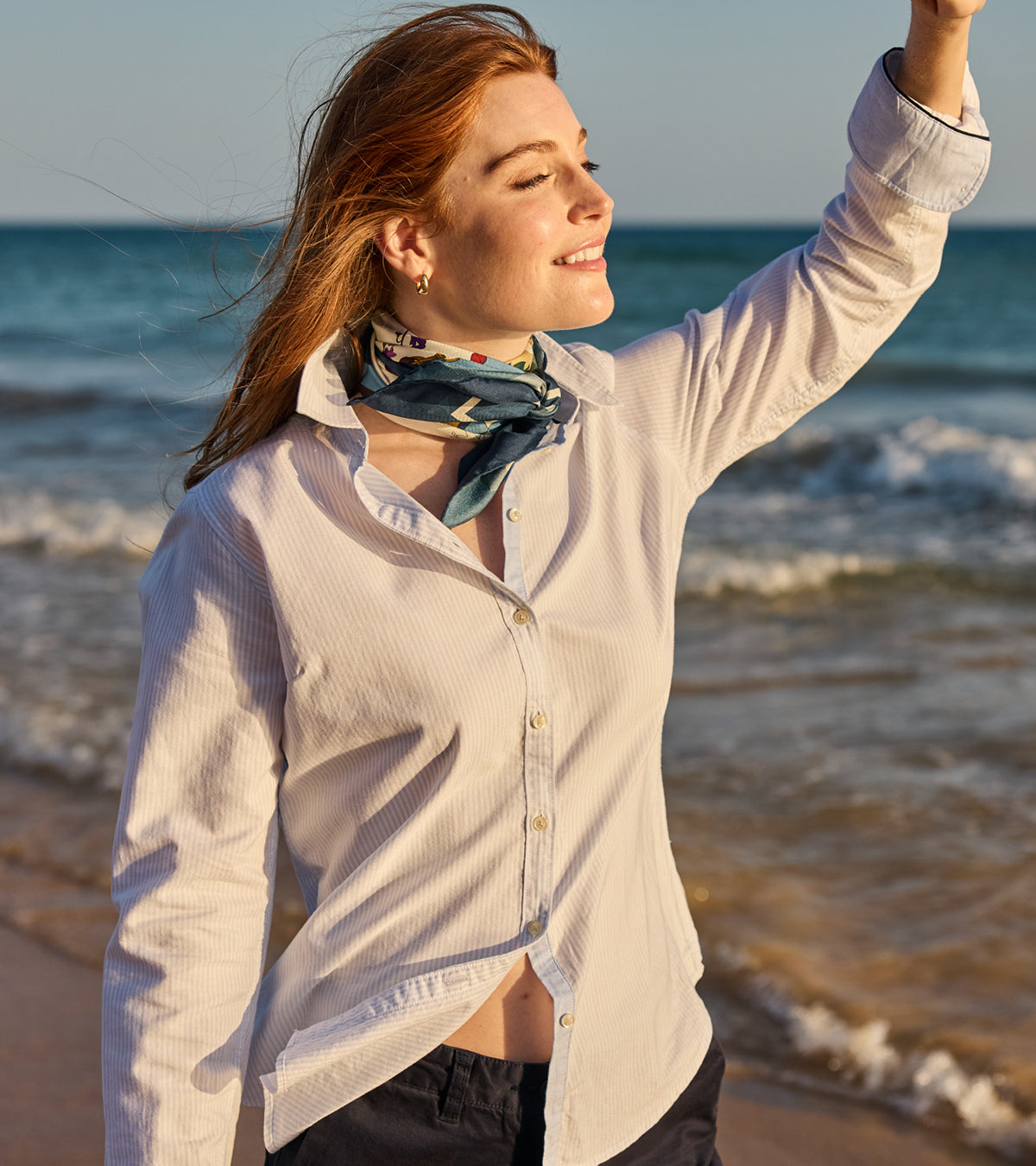 A woman on a beach wearing a light beige button-up shirt and a blue patterned neck scarf, with ocean waves and a clear sky in the background.