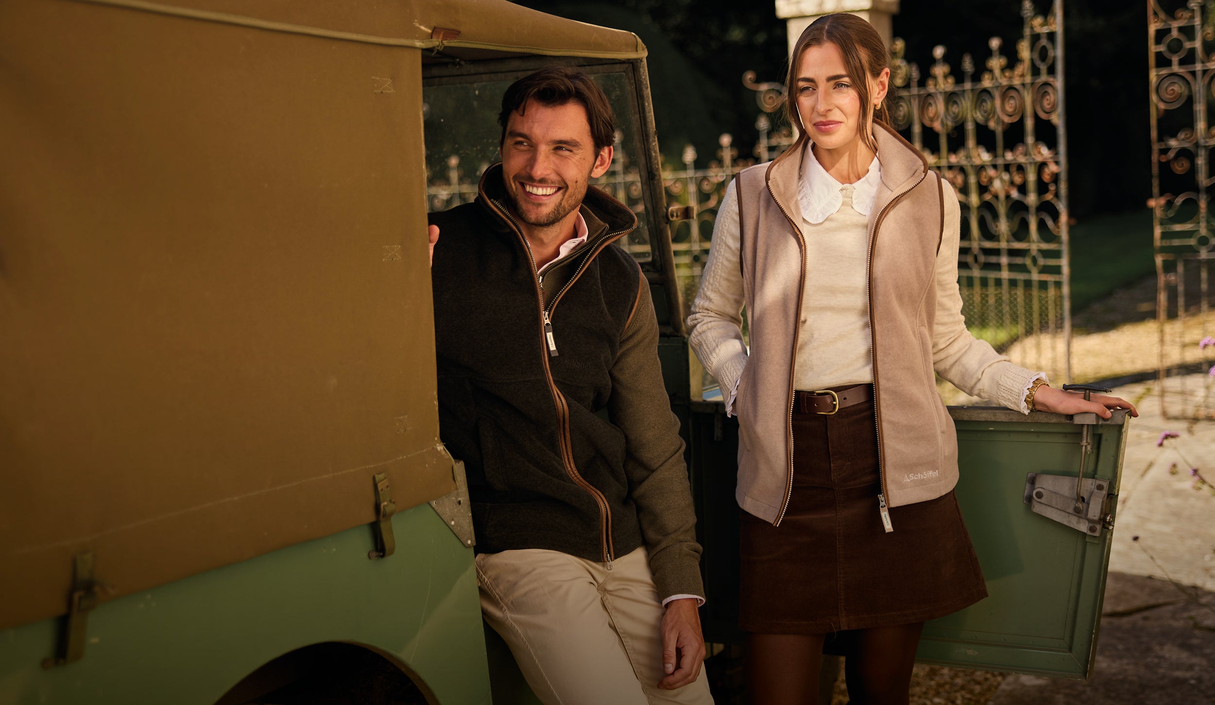 A man and a woman standing beside a vintage green vehicle at night, with the man wearing a dark zip-up gilet and light trousers and the woman wearing a beige gilet over a white top and brown skirt.