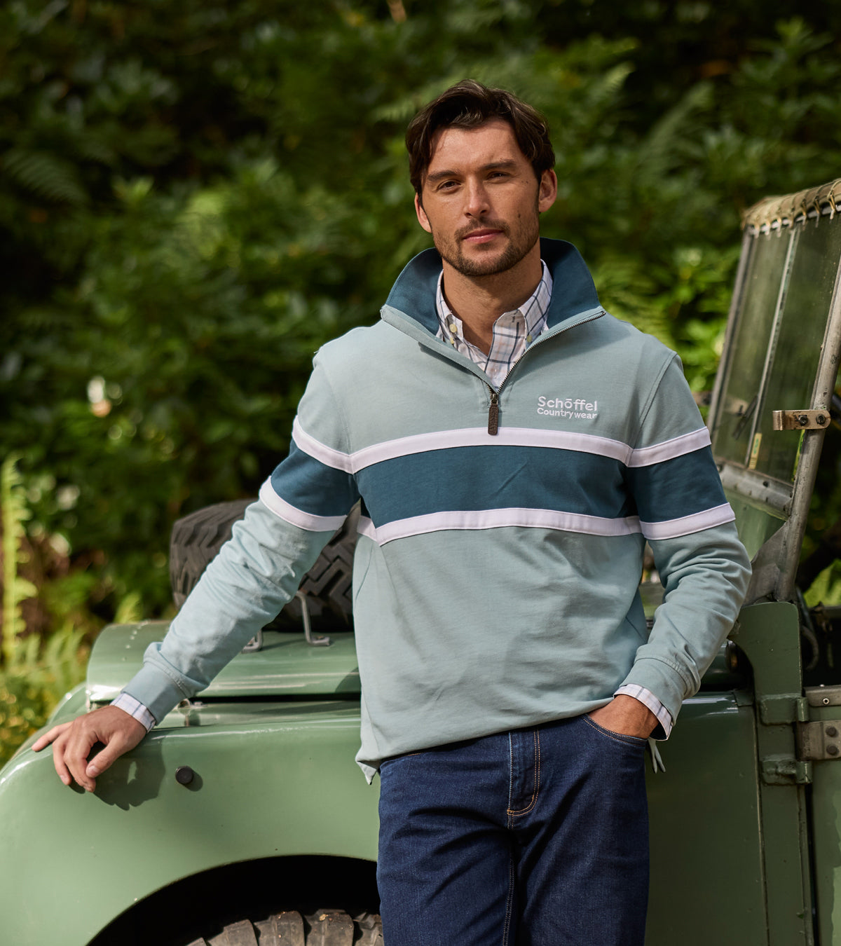 Man wearing a light blue jacket with white stripes standing next to a green vehicle in a forest setting
