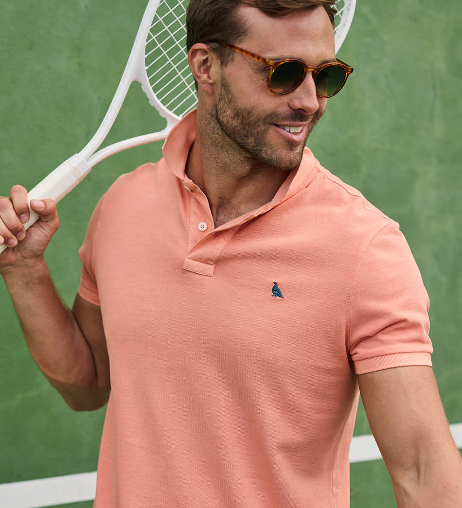 Man wearing a pink polo shirt with a logo, holding a tennis racket on a tennis court.