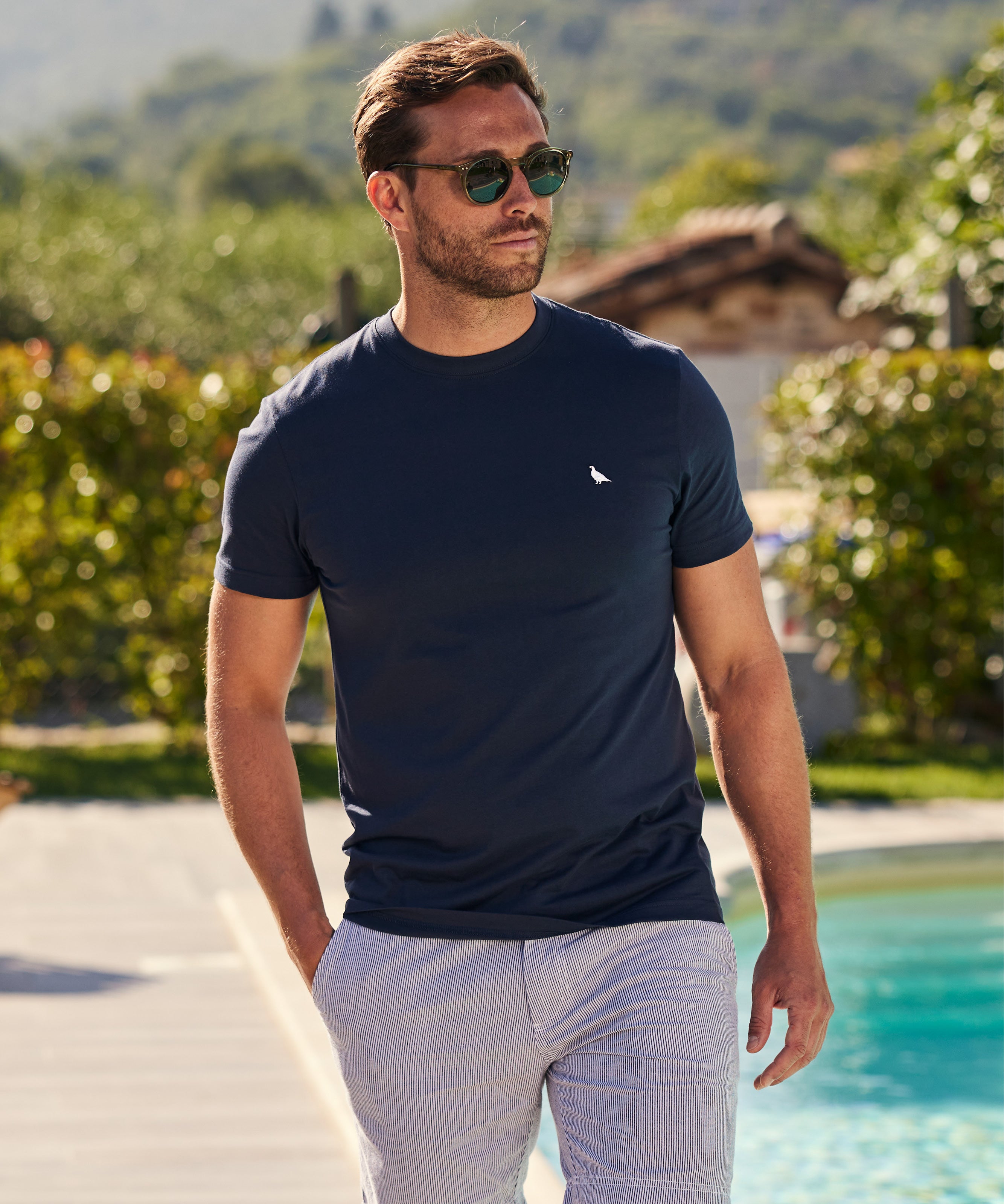 Man wearing a navy blue t-shirt and striped swim shorts, walking poolside on a sunny day with sunglasses on and a relaxed, confident expression.