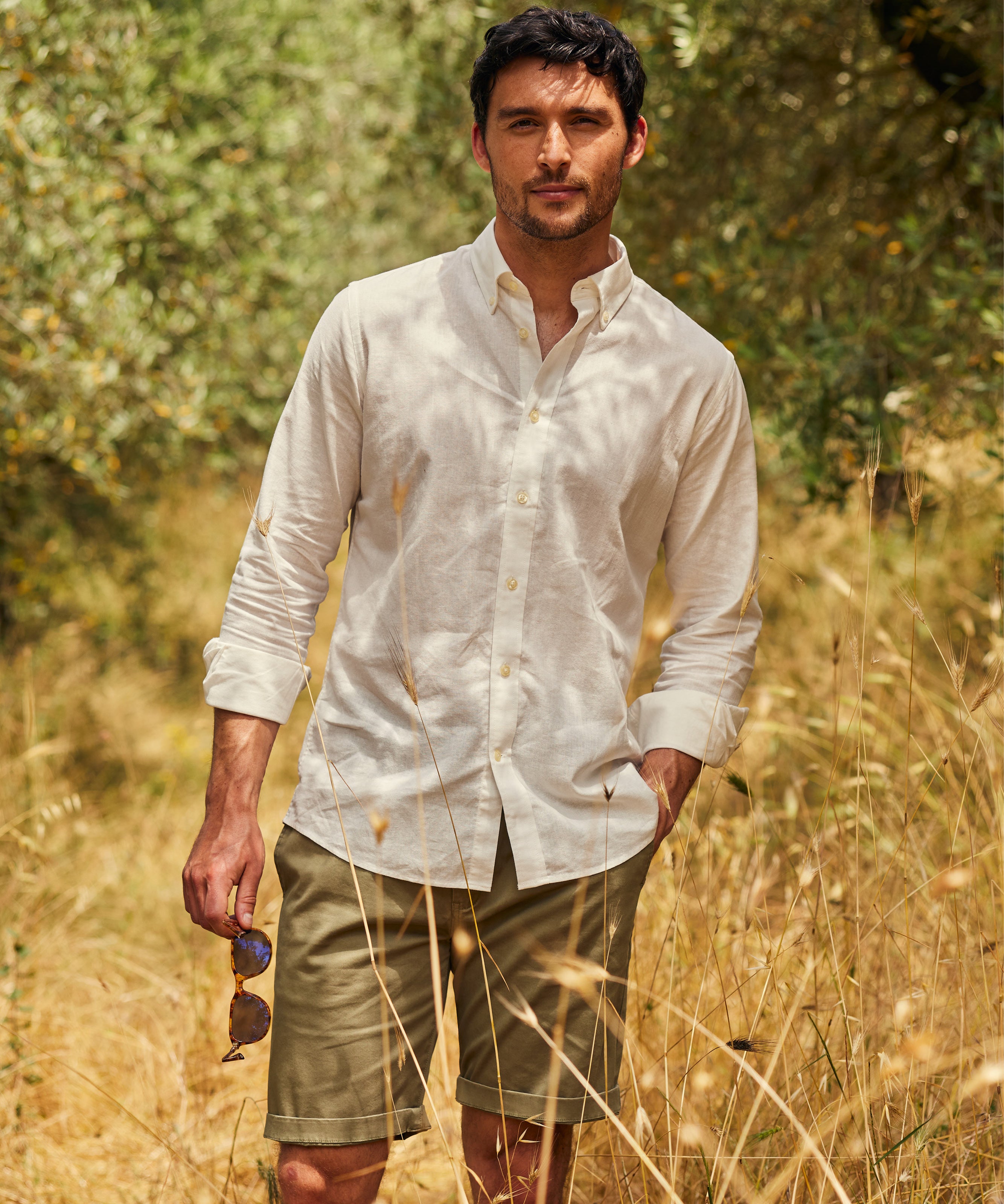 Man standing in a sunlit field wearing a white button-down shirt and olive green shorts, holding sunglasses in one hand, surrounded by tall grass and greenery.