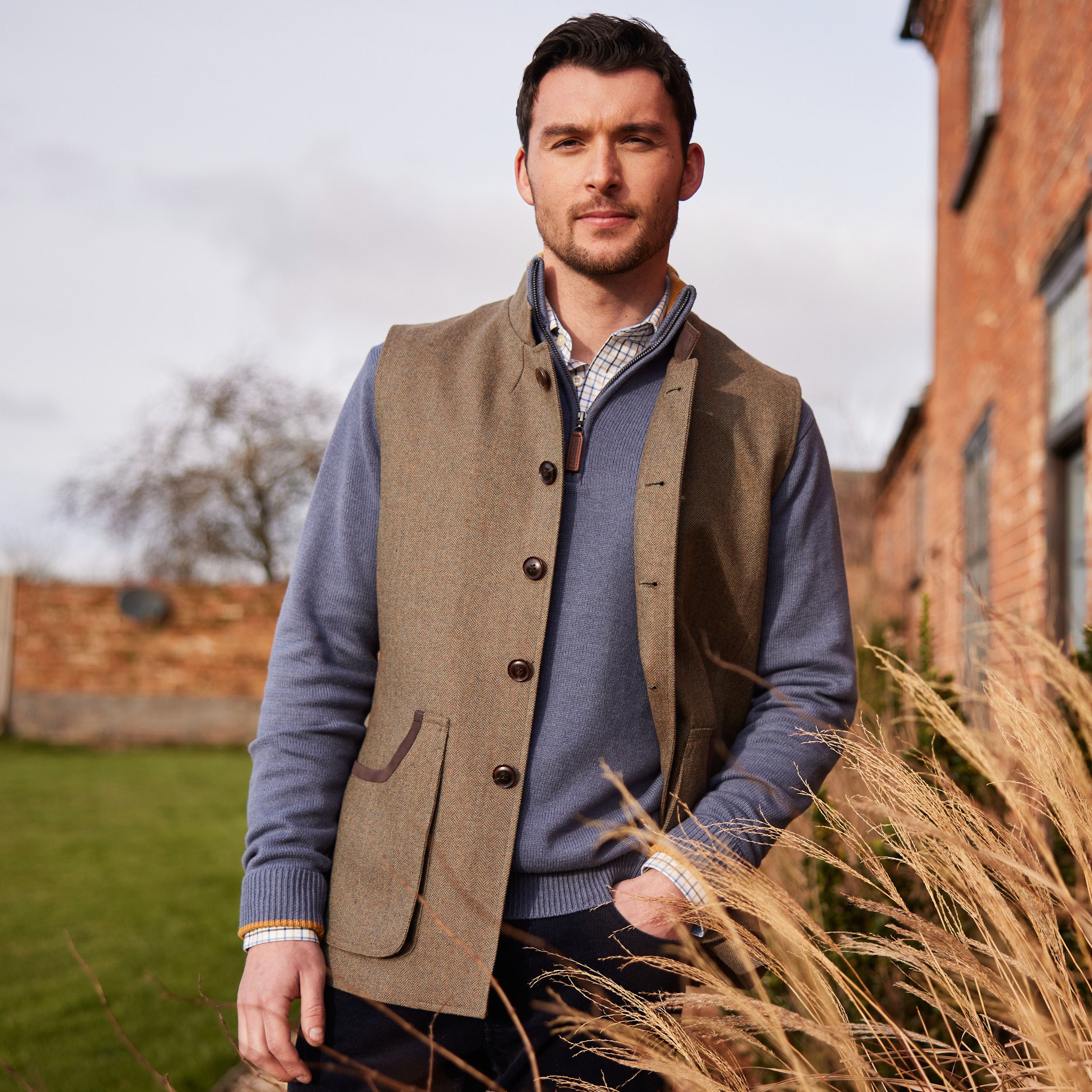 A man wearing a tweed waistcoat with blue quarter zip jumper and checked shirt. It is paired with dark navy jeans and he is standing outside a red brick building on a grass lawn.