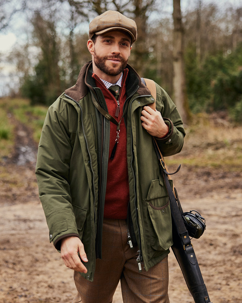 A man wearing a red knitted jumper, green country jacket and tweed trousers. He has a gun on his shoulder and he is looking away from the camera.