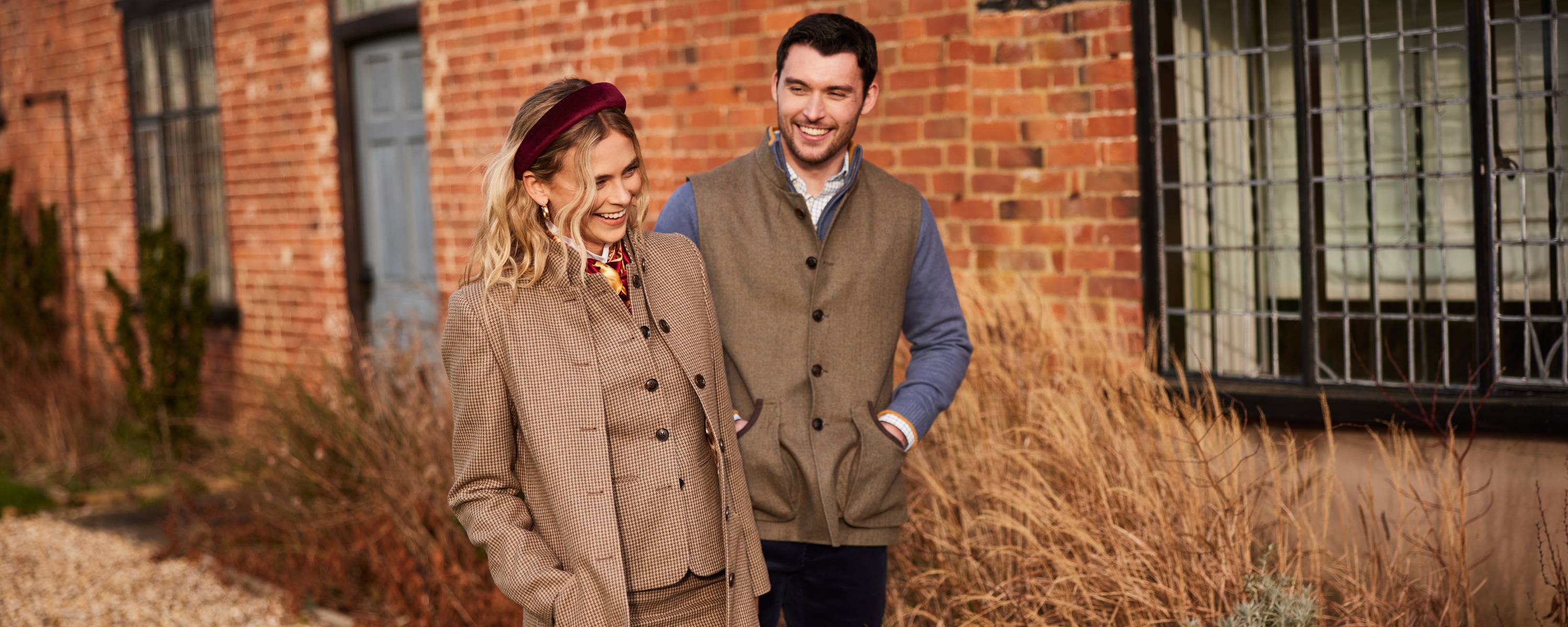 A stylishly dressed man and woman walk together outdoors, smiling and laughing. The woman, wearing a brown tweed jacket, matching waistcoat, and a burgundy velvet headband, has wavy blonde hair and a patterned scarf around her neck. The man, slightly behind her, is wearing a brown tweed gilet over a blue shirt with a relaxed expression. They are walking past a rustic red brick building with large windows, surrounded by dried grasses and shrubs, giving an autumnal countryside feel.
