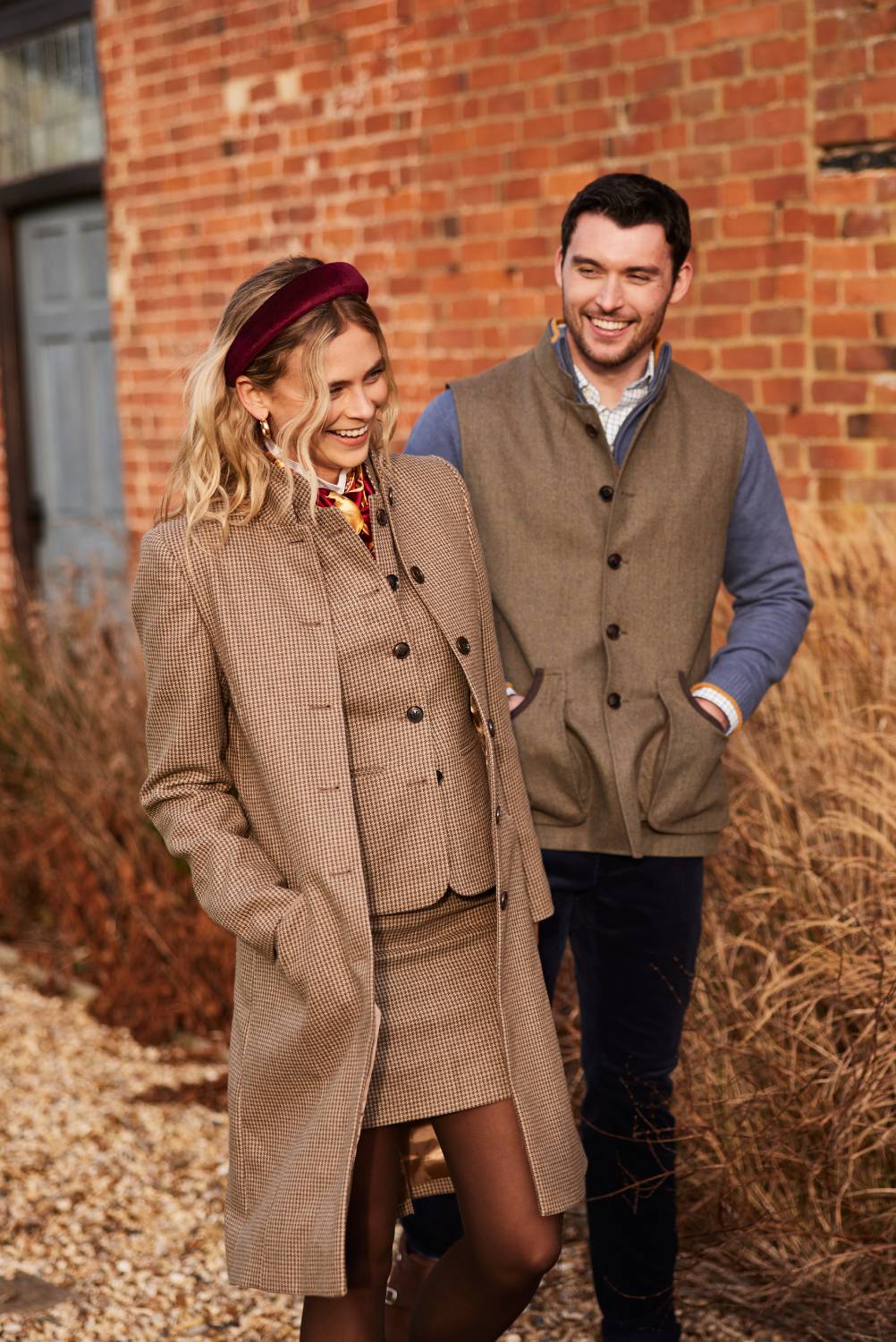 A stylishly dressed man and woman walk together outdoors, smiling and laughing. The woman, wearing a brown tweed jacket, matching waistcoat, and a burgundy velvet headband, has wavy blonde hair and a patterned scarf around her neck. The man, slightly behind her, is wearing a brown tweed gilet over a blue shirt with a relaxed expression. They are walking past a rustic red brick building with large windows, surrounded by dried grasses and shrubs, giving an autumnal countryside feel.