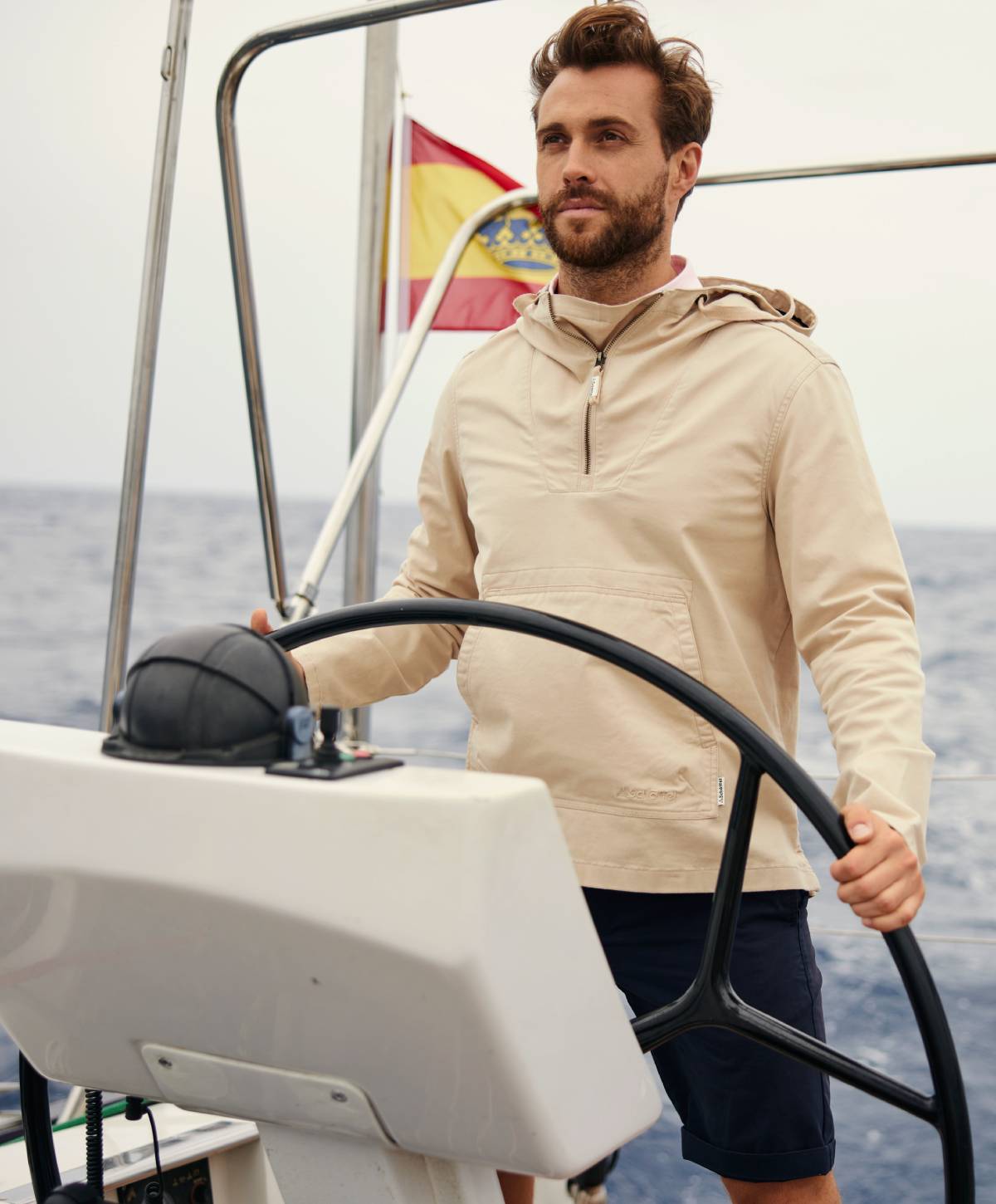 A man standing on a boat in a beige jacket and navy shorts.