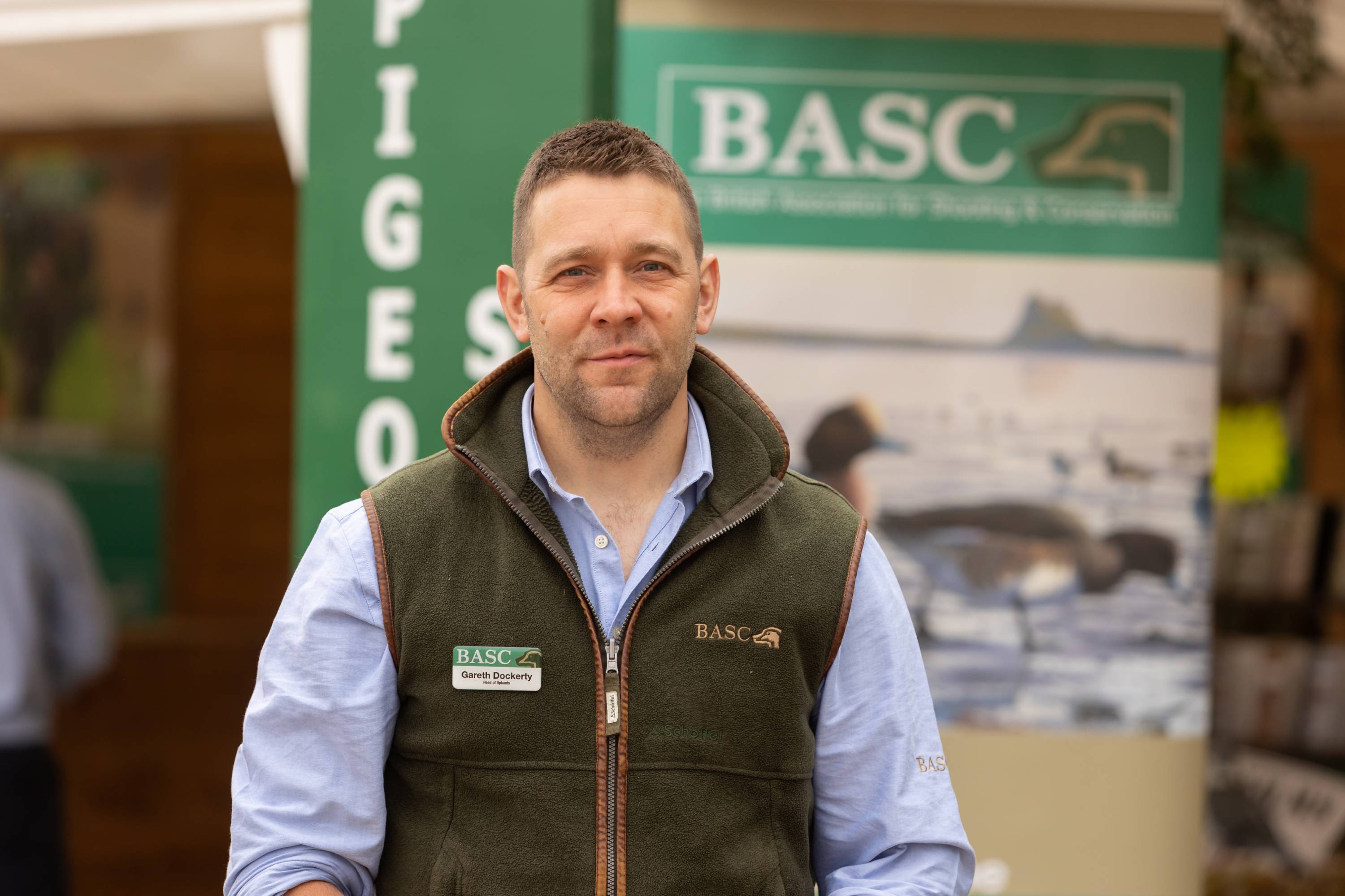 A man wearing a blue shirt and green gilet, standing in front of a sign, looking towards the camera.