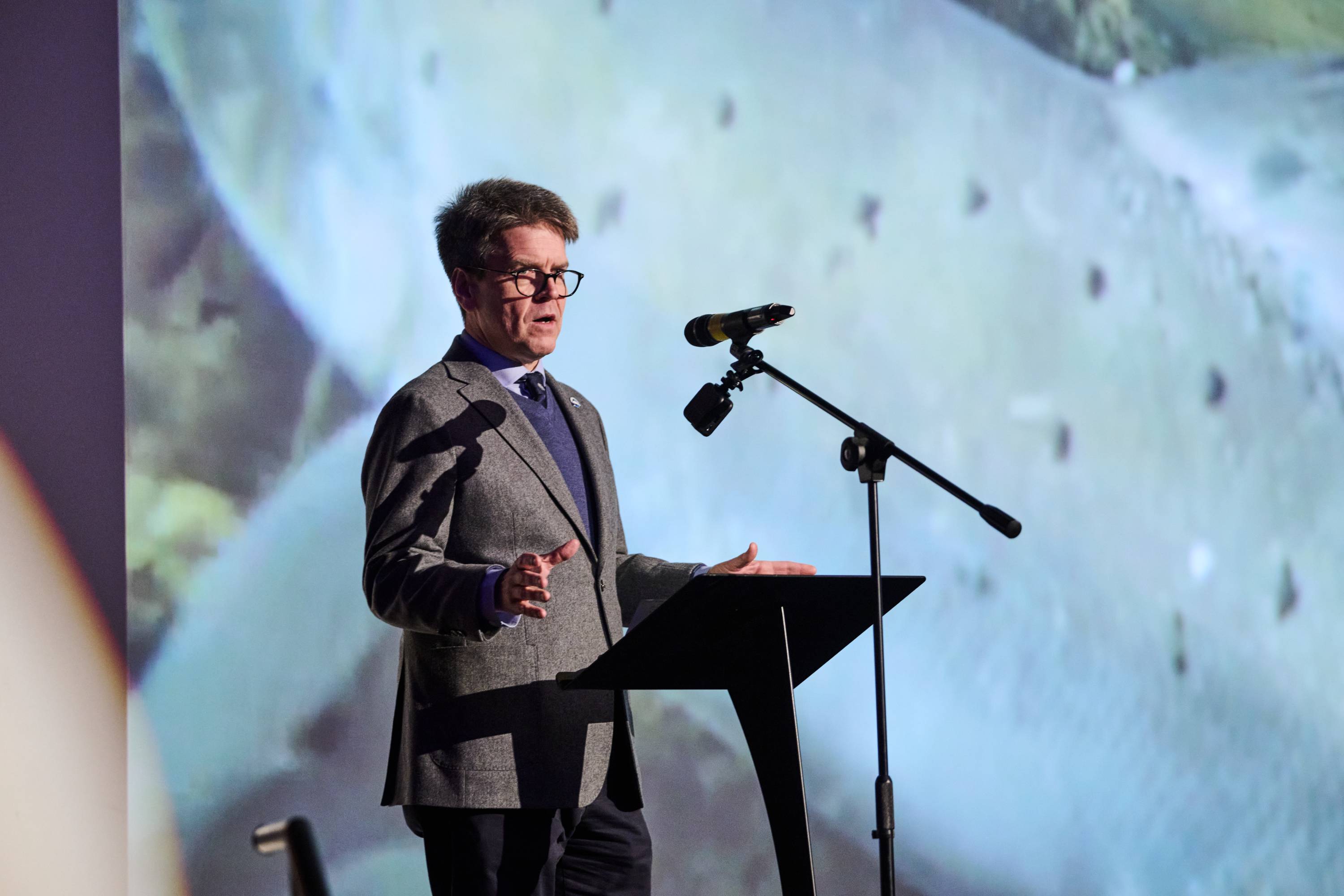A man wearing a smart outfit, standing on a stage in front of a large screen and microphone giving a presentation.