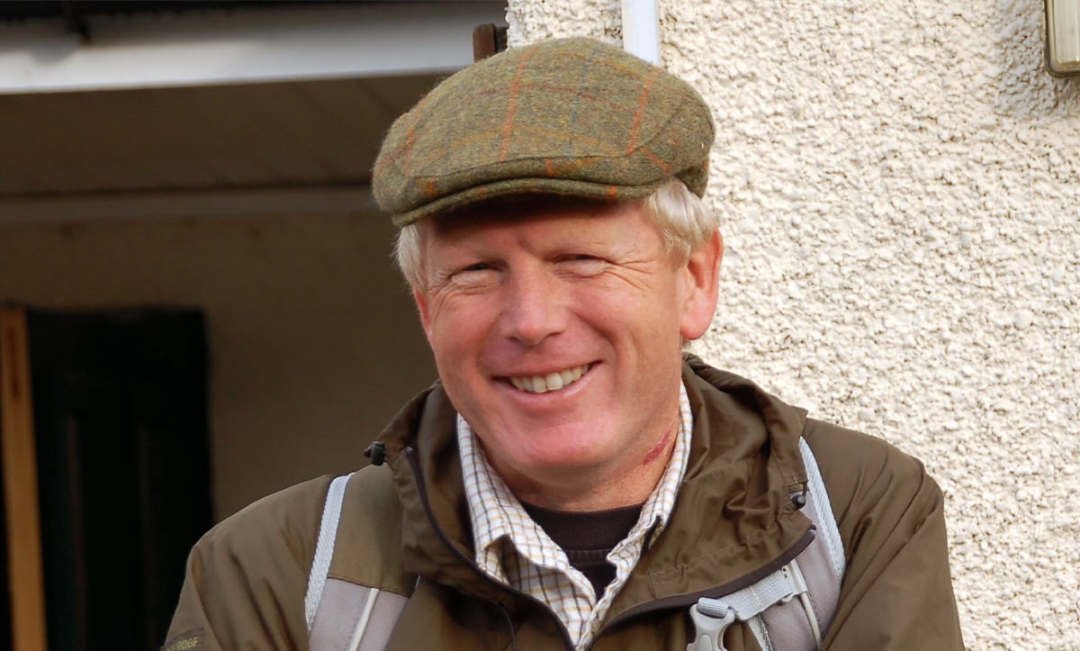 A man smiling, wearing a tweed cap, checked shirt and brown coat.