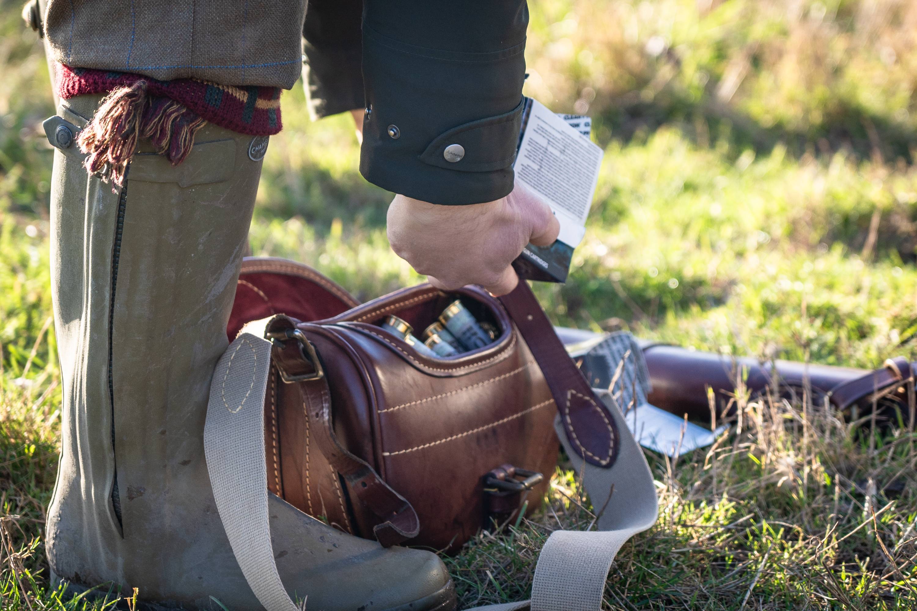 A person looking into an ammo bag wearing tweed culottes, a green jacket and green wellingtons.