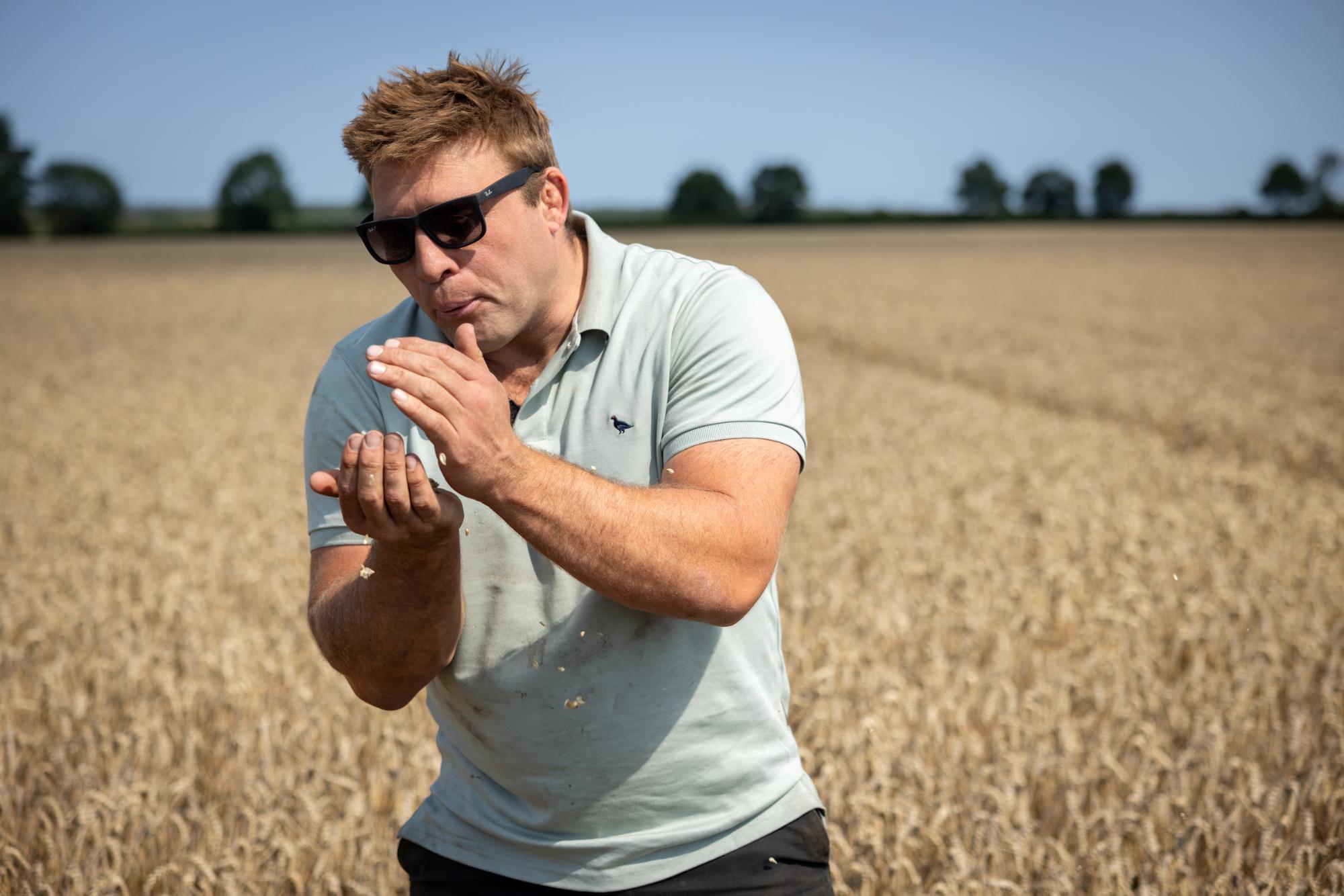 Man in sunglasses and light polo shirt standing in a field, blowing on his hands