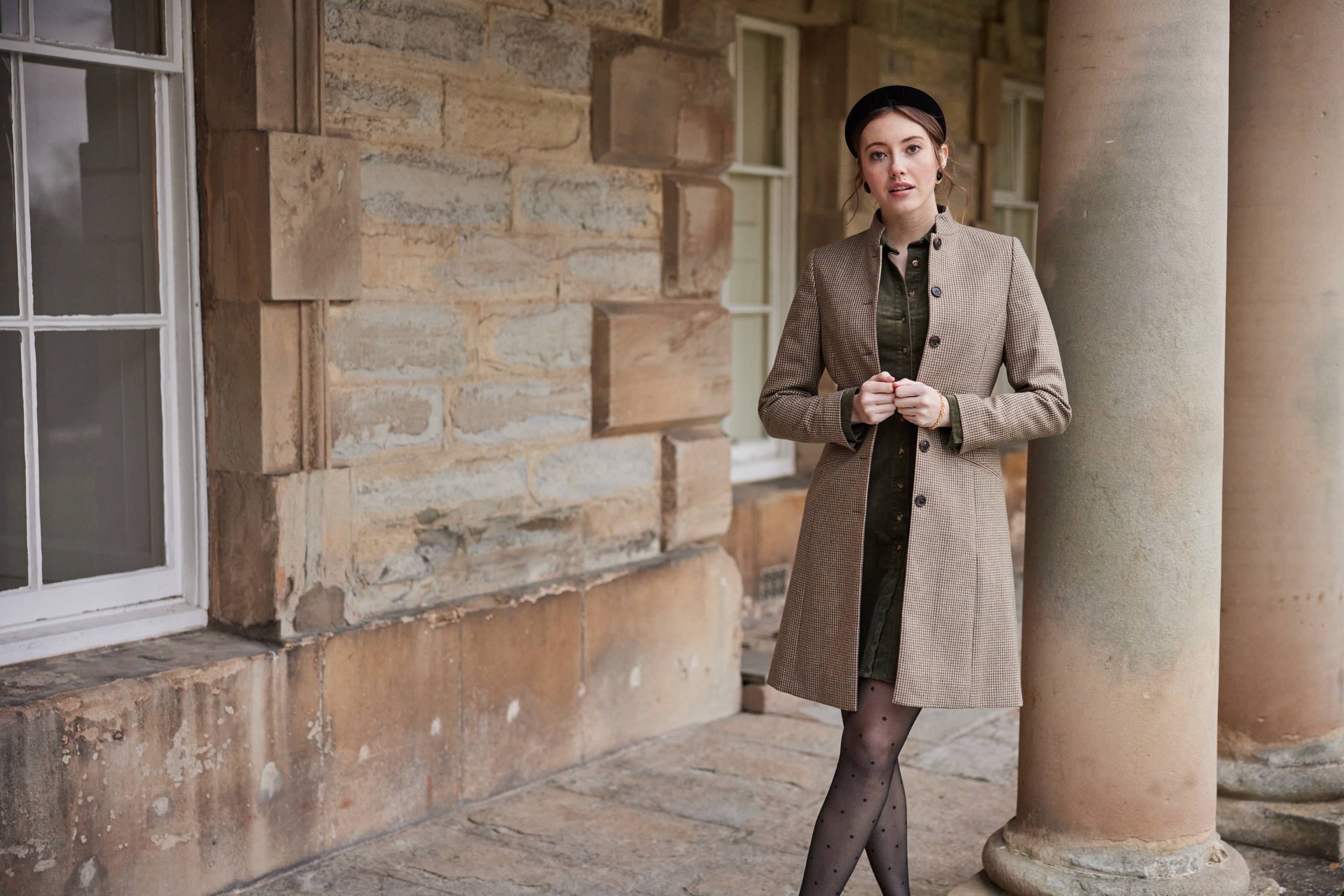 A lady standing in front of a stone building wearing a green dress and tweed jacket.