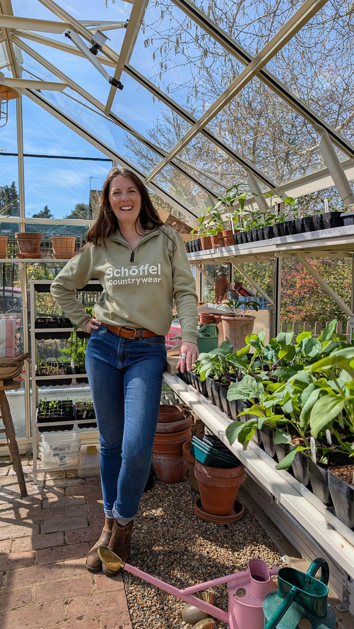 Woman standing in a greenhouse with plants and gardening tools.