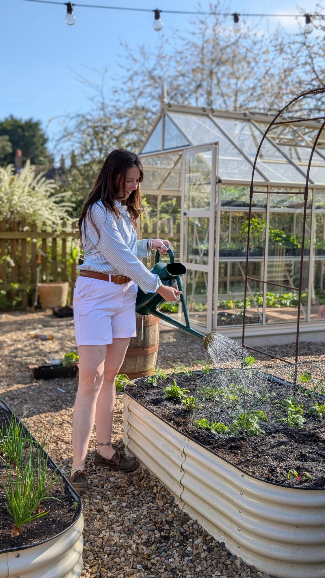 Woman watering plants in a garden with a greenhouse in the background