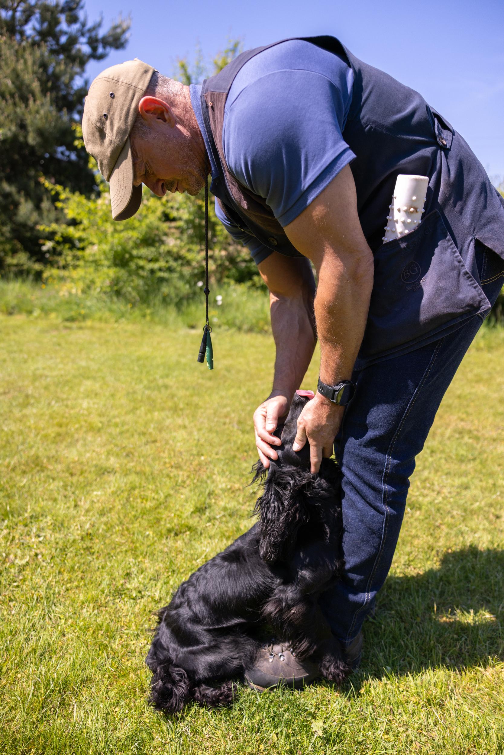 Man petting a black dog on a grassy field with trees in the background
