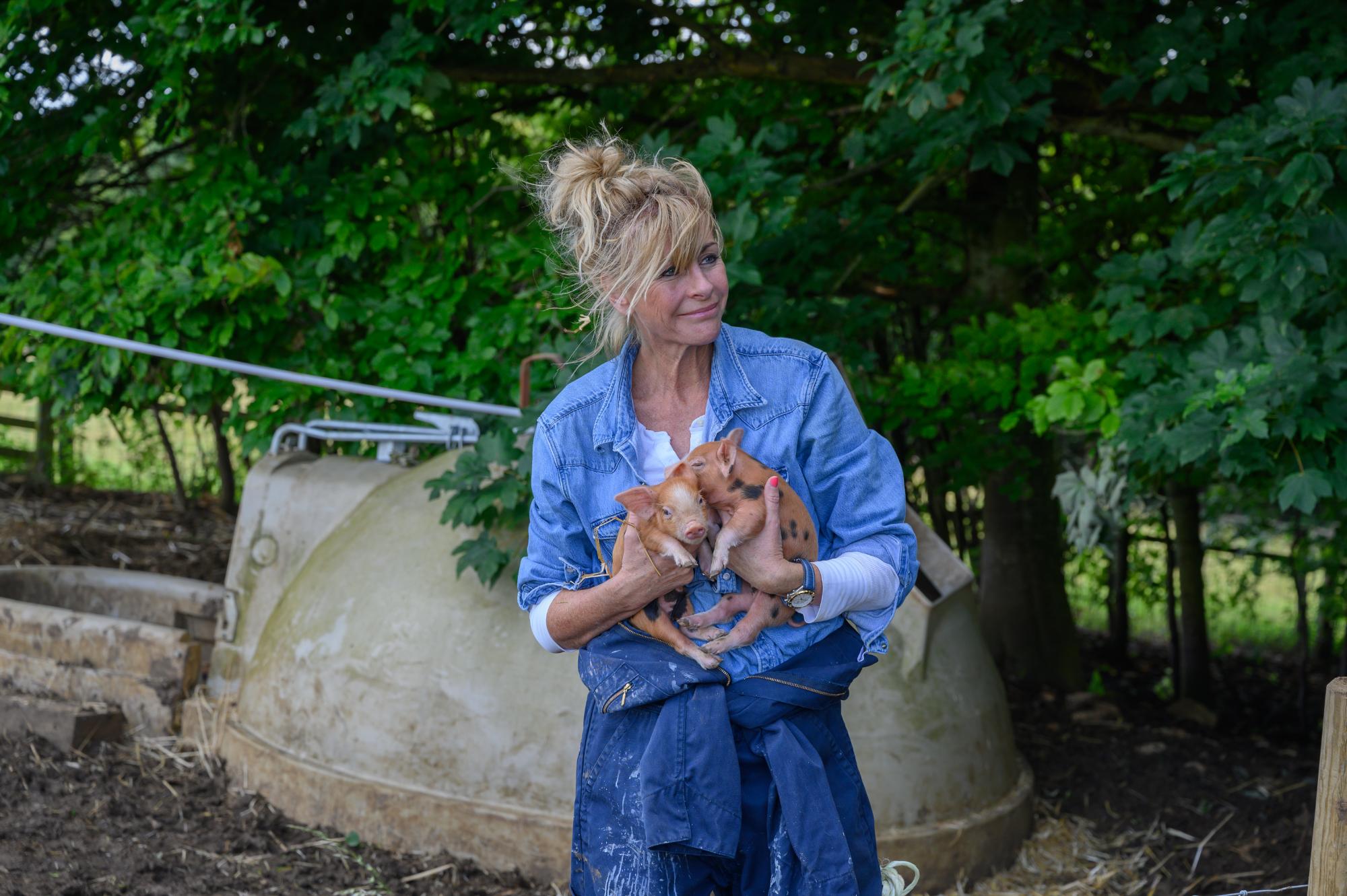 Woman holding piglets in a rural setting with trees and a water tank in the background.