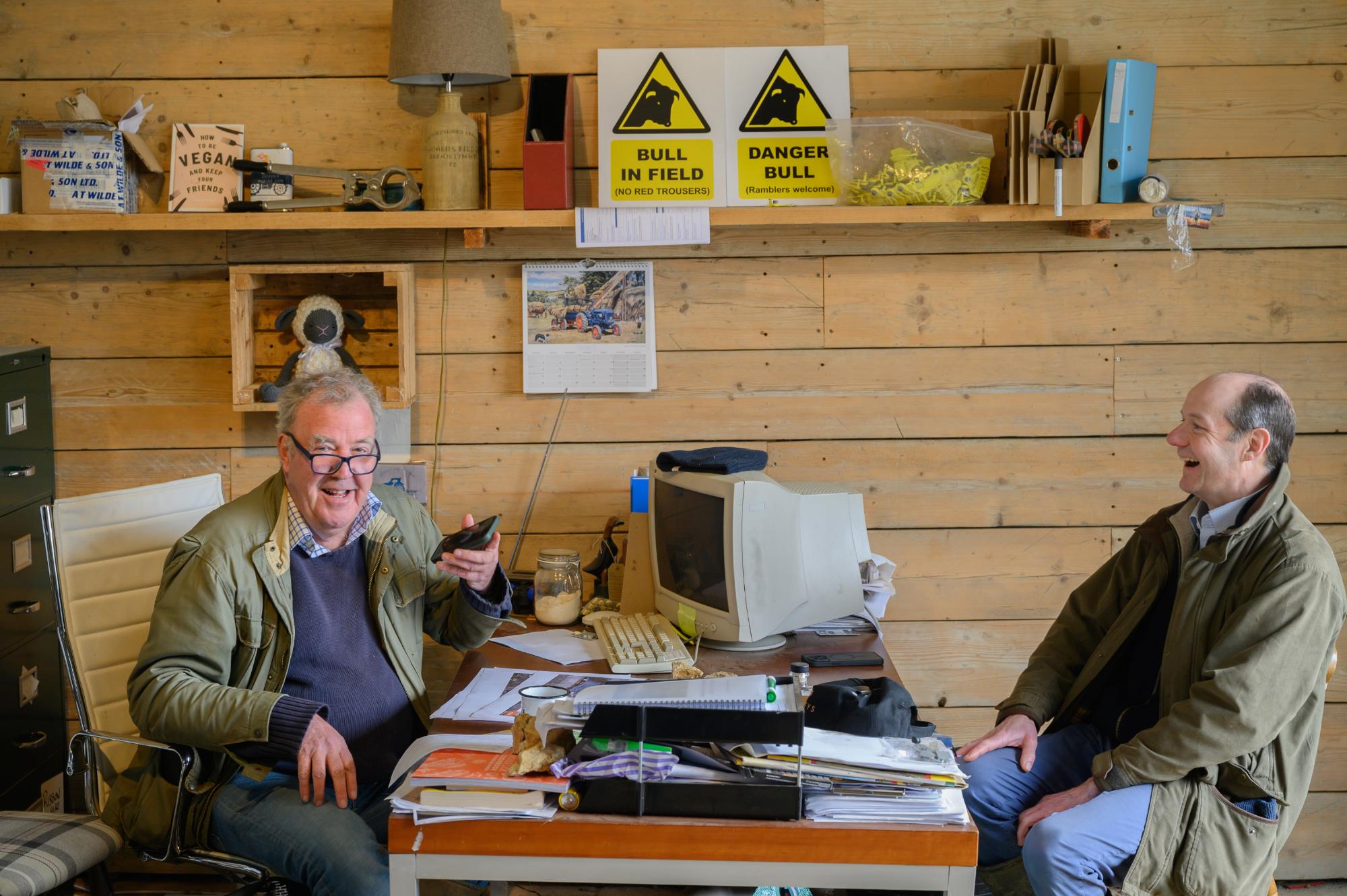 Two men sitting at a desk in a wooden room with shelves and warning signs.