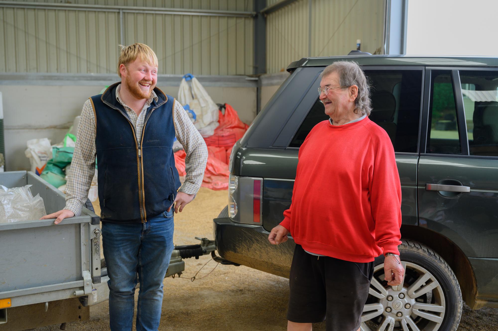Two men standing next to a vehicle in an indoor setting with various items around.