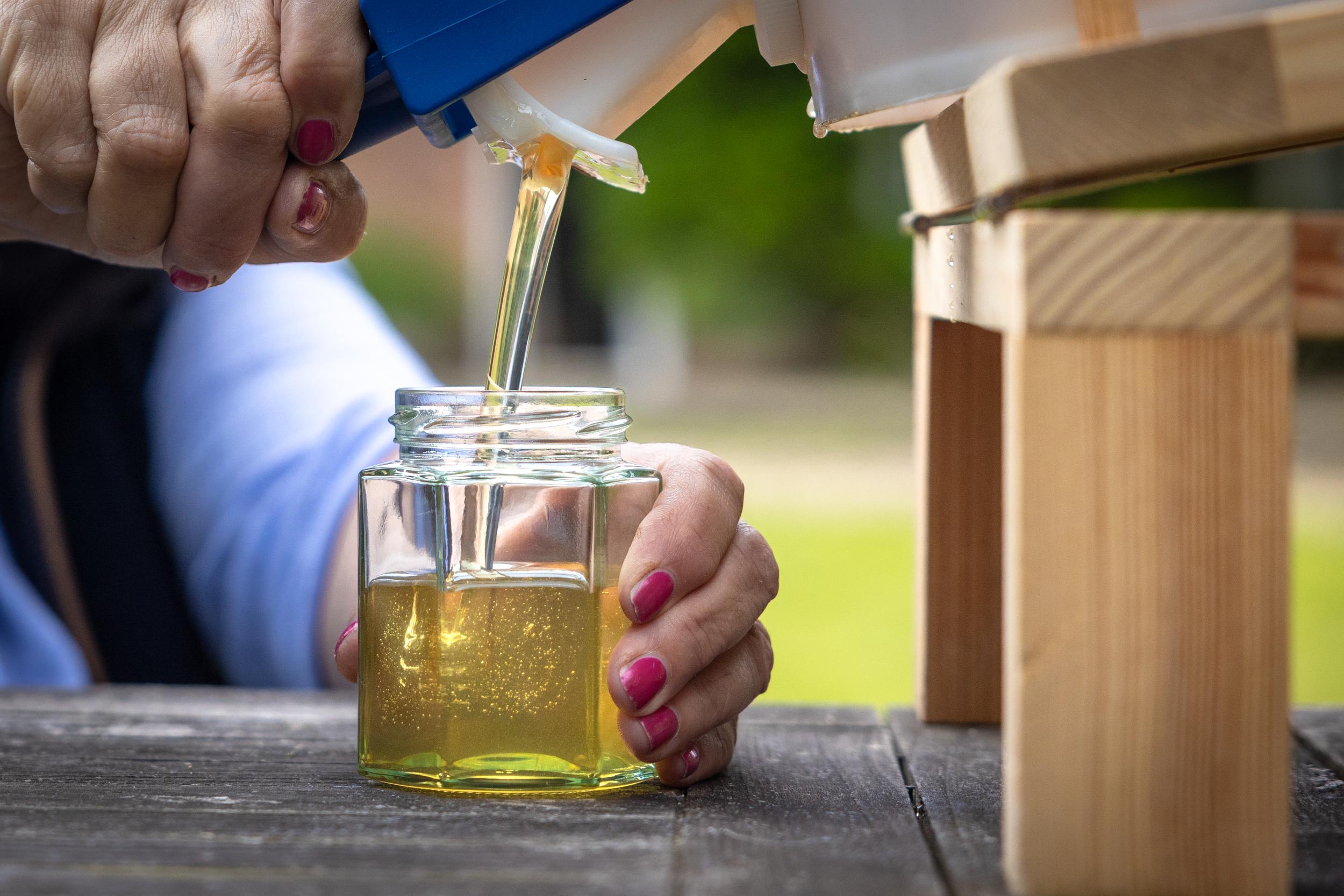 Hand pouring honey from a bottle into a glass jar on a wooden surface.
