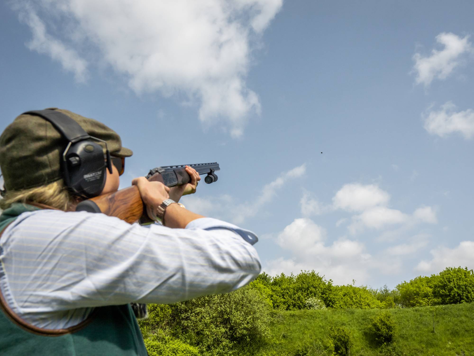 Person shooting a gun in an outdoor setting with trees and blue sky.