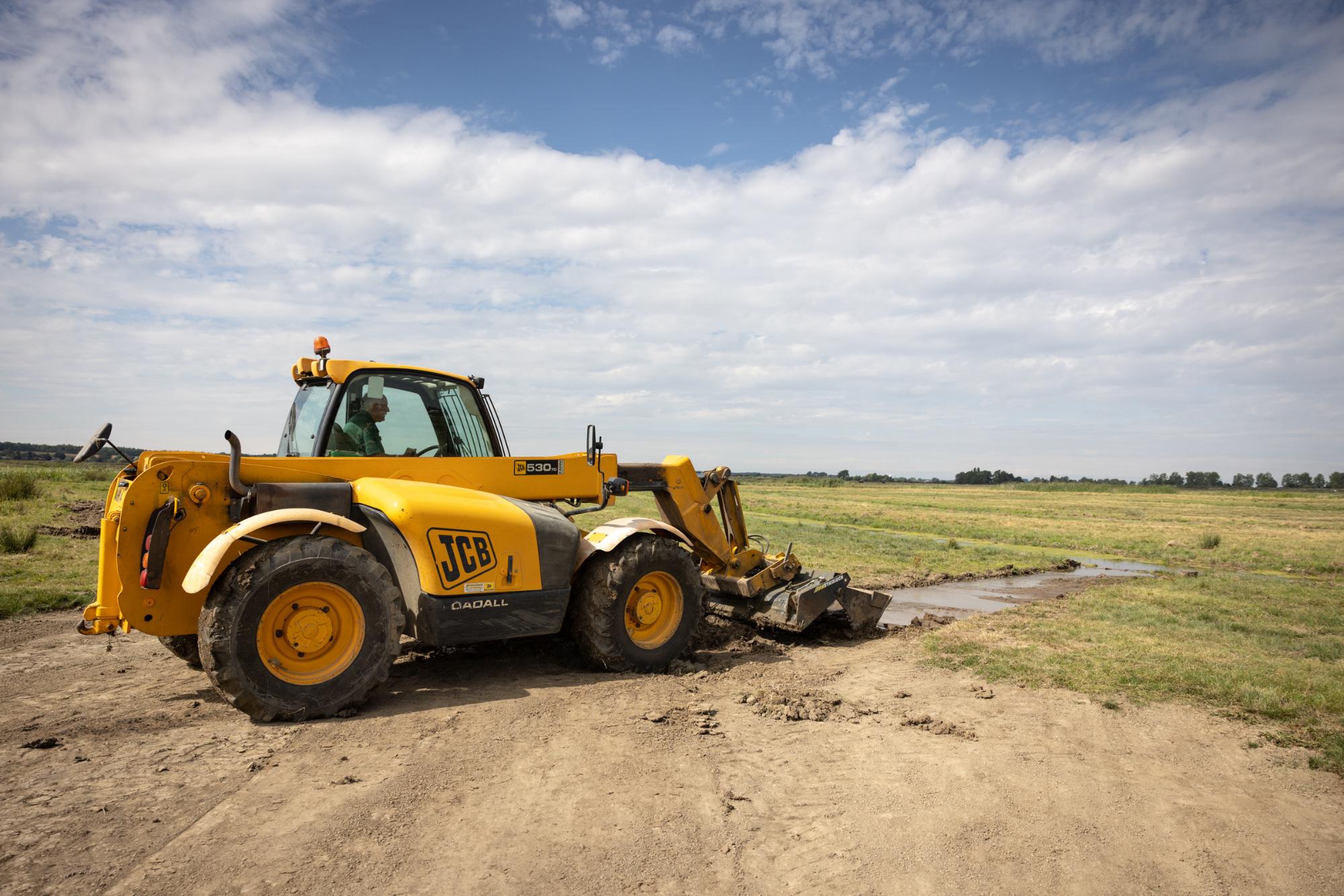 Yellow JCB excavator working on a dirt path with a grassy field and blue sky in the background.